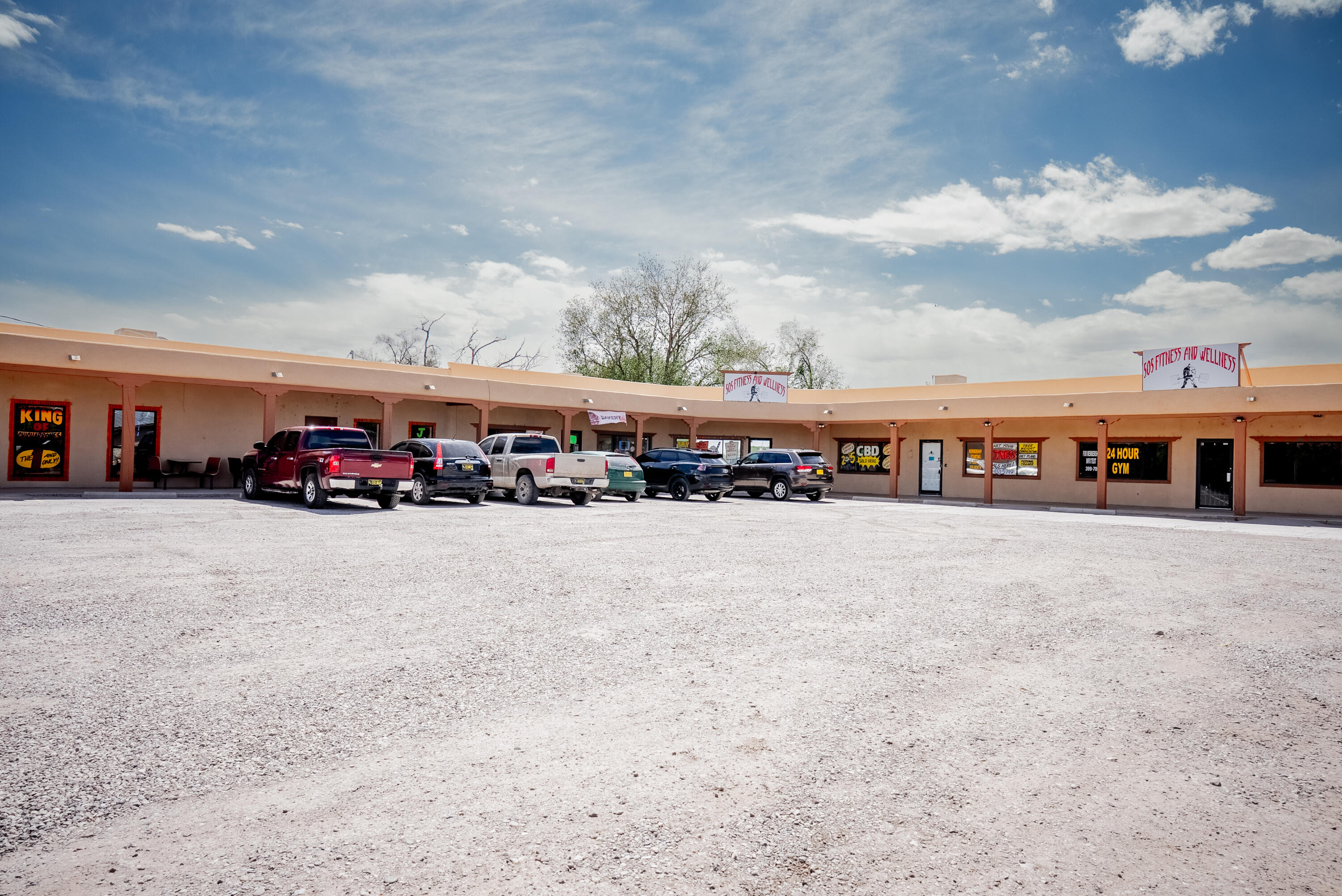Commercial strip with fitness businesses and parked vehicles along a broad gravel lot.