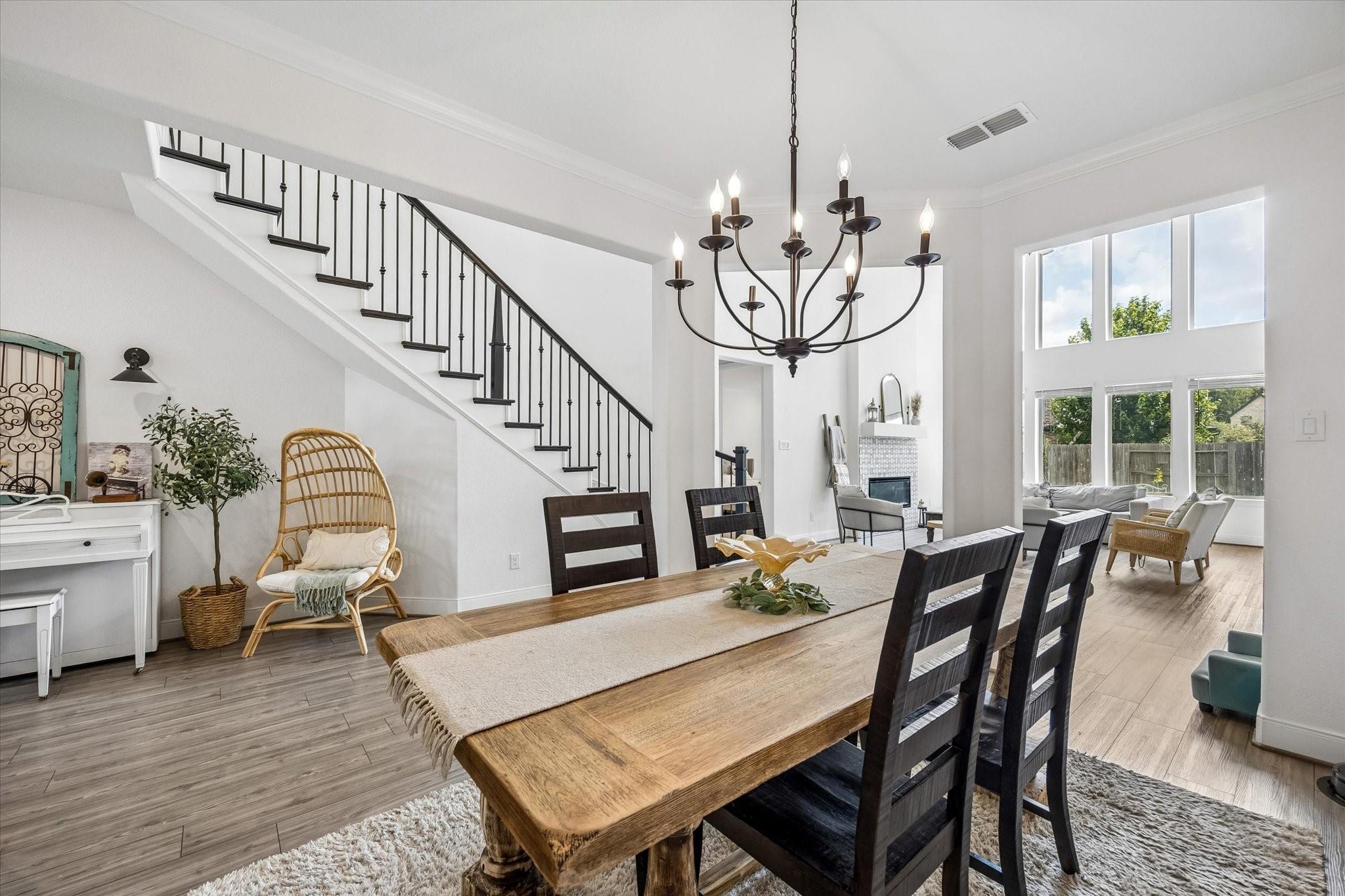 Dining room with a rustic wood table, black chandelier, staircase, and tall windows opening to the living area.