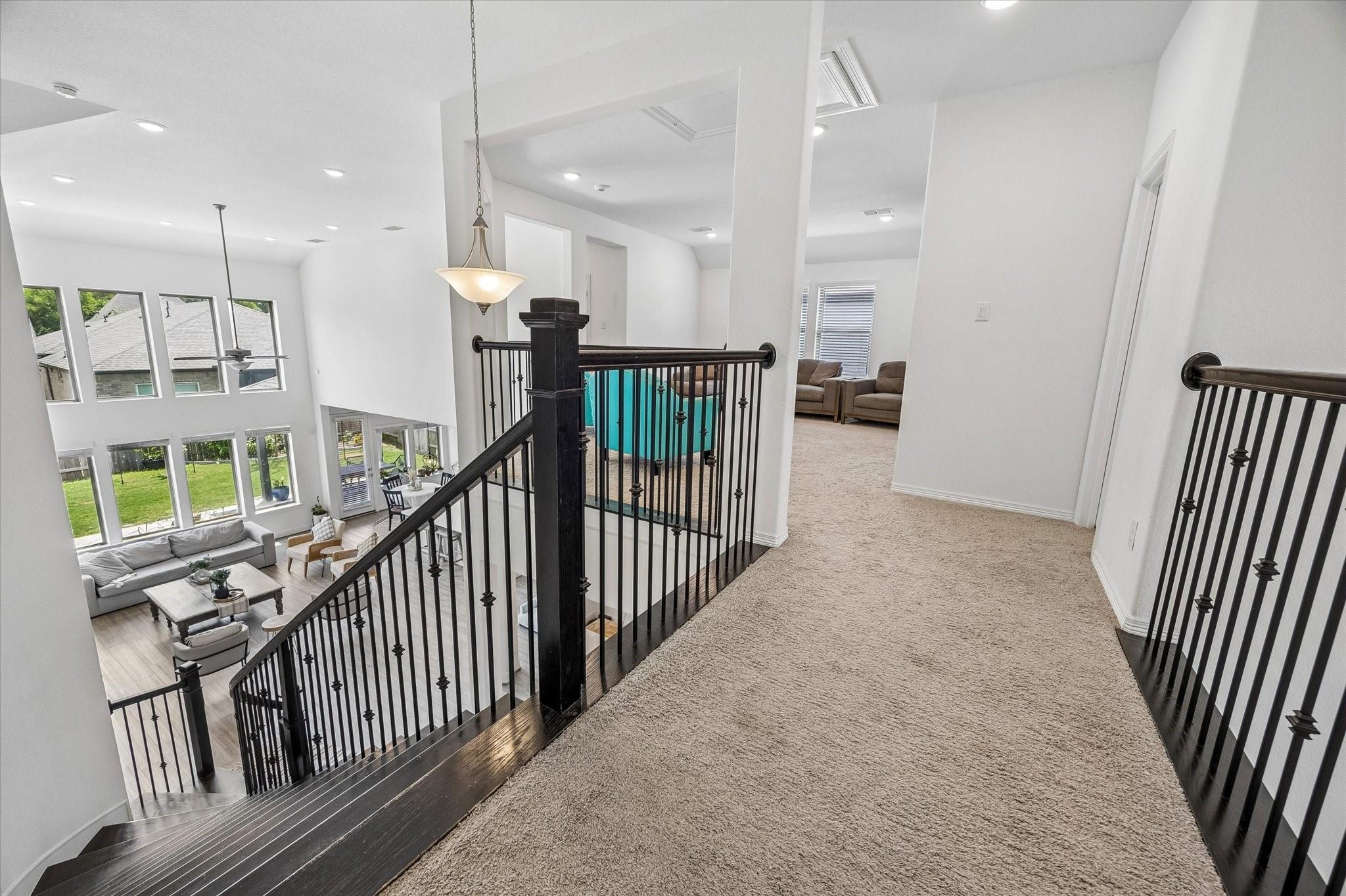 Upstairs landing with black iron railings overlooking a bright living room through double-height windows.