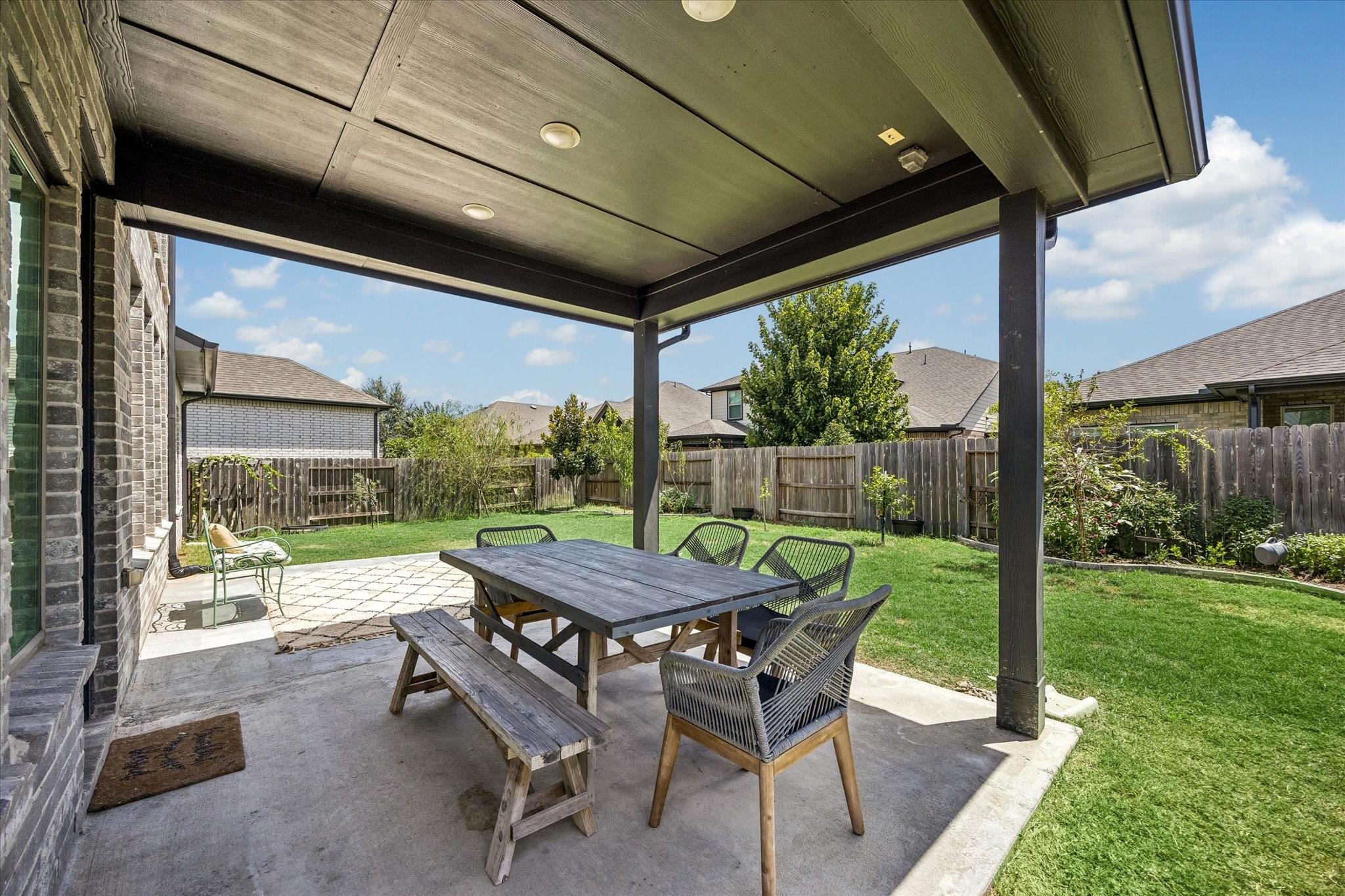 Covered patio with outdoor dining table and chairs beside a fenced backyard lawn.
