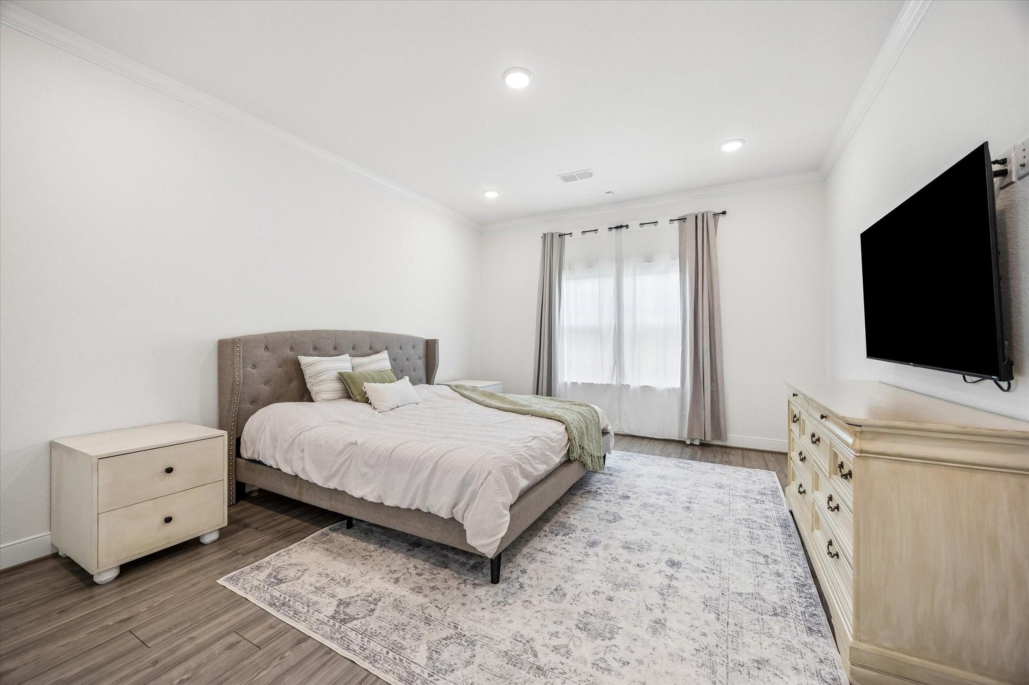 Bedroom with a gray tufted bed, pale area rug, and wall-mounted television.