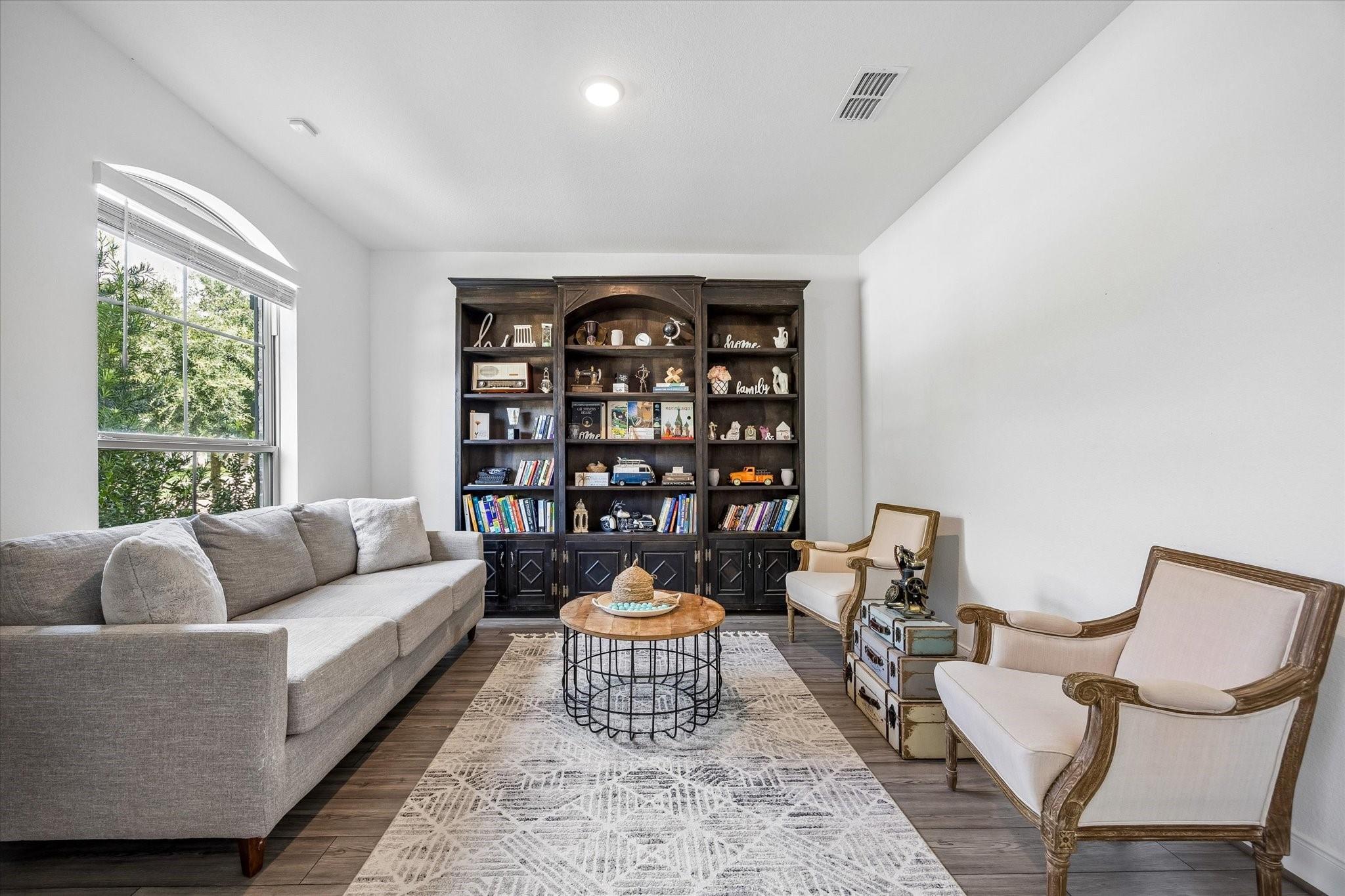 Sitting room with a dark built-in bookcase, gray sofa, cream armchairs, and arched window.