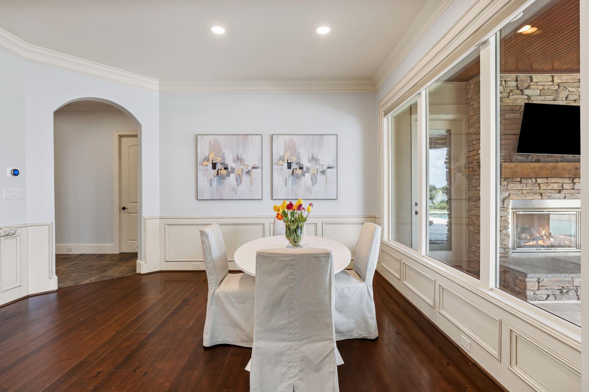 Dining area with a round table beside tall windows overlooking a stone fireplace.