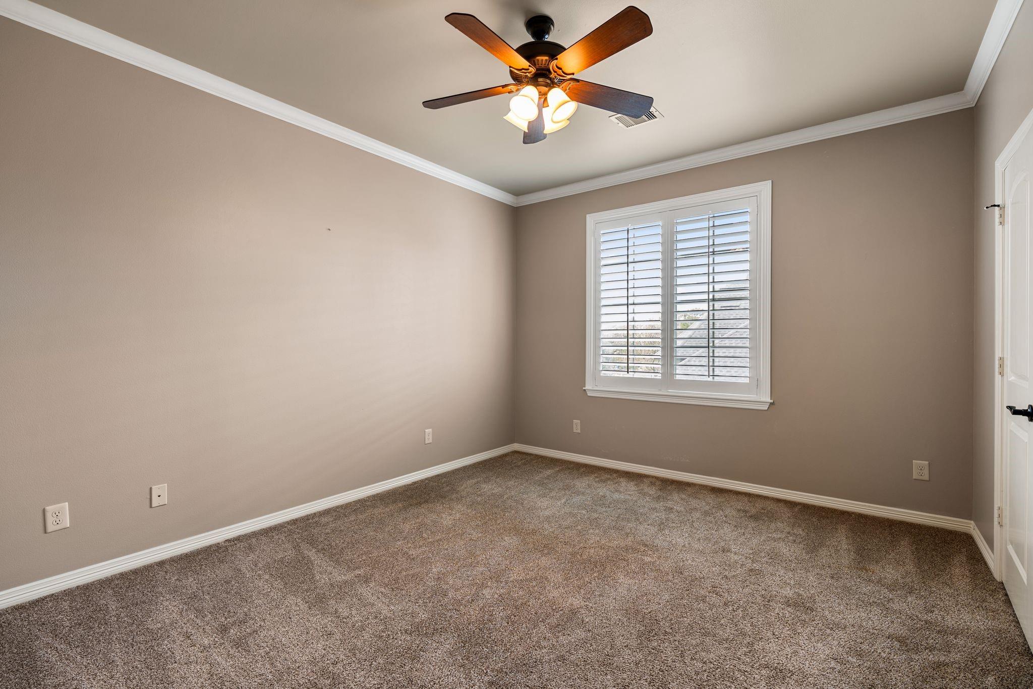 Empty carpeted bedroom with a lit ceiling fan and white plantation shutters.