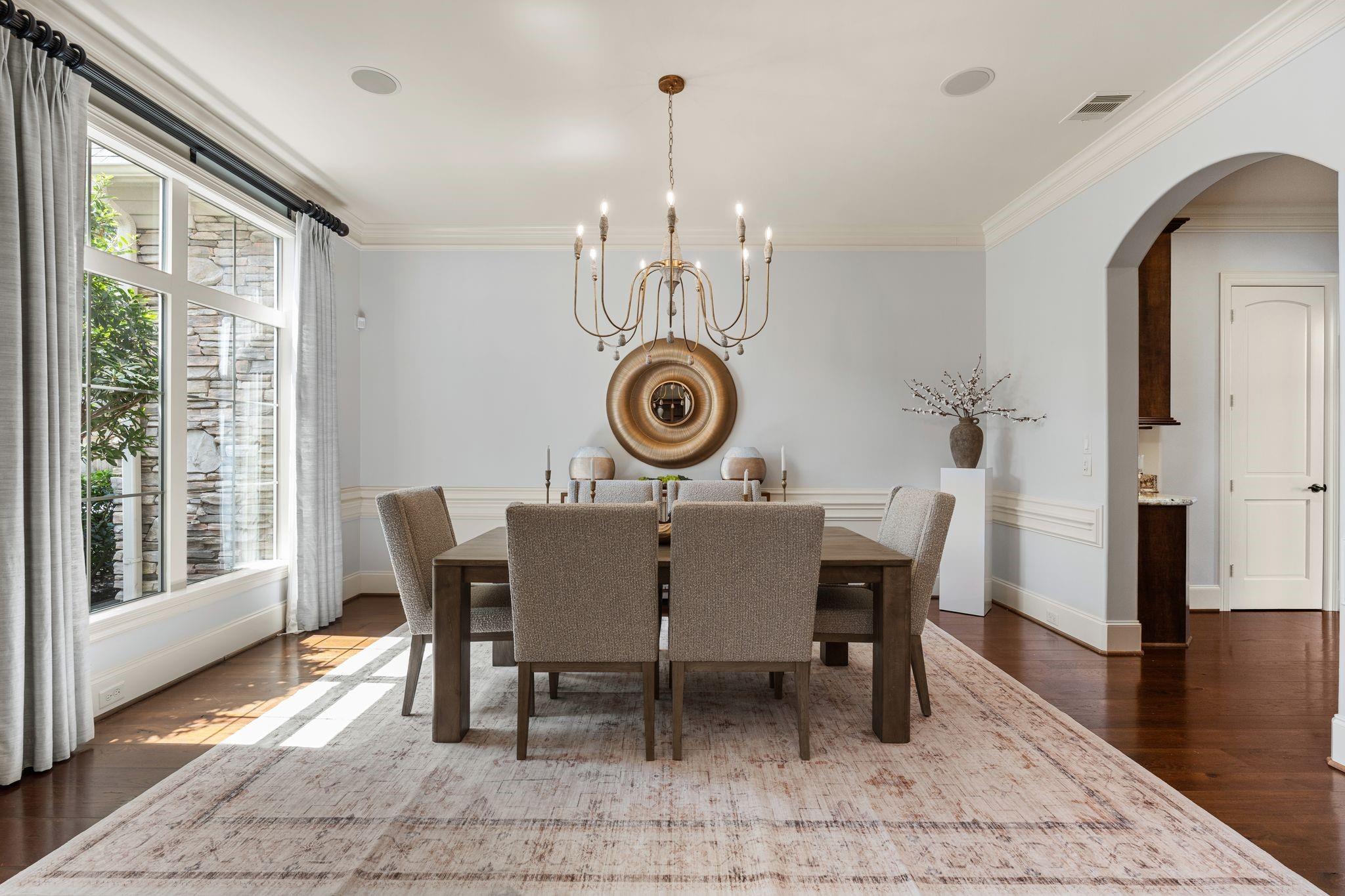 Dining room with a dark wood table, upholstered chairs, brass chandelier, and tall curtained windows.