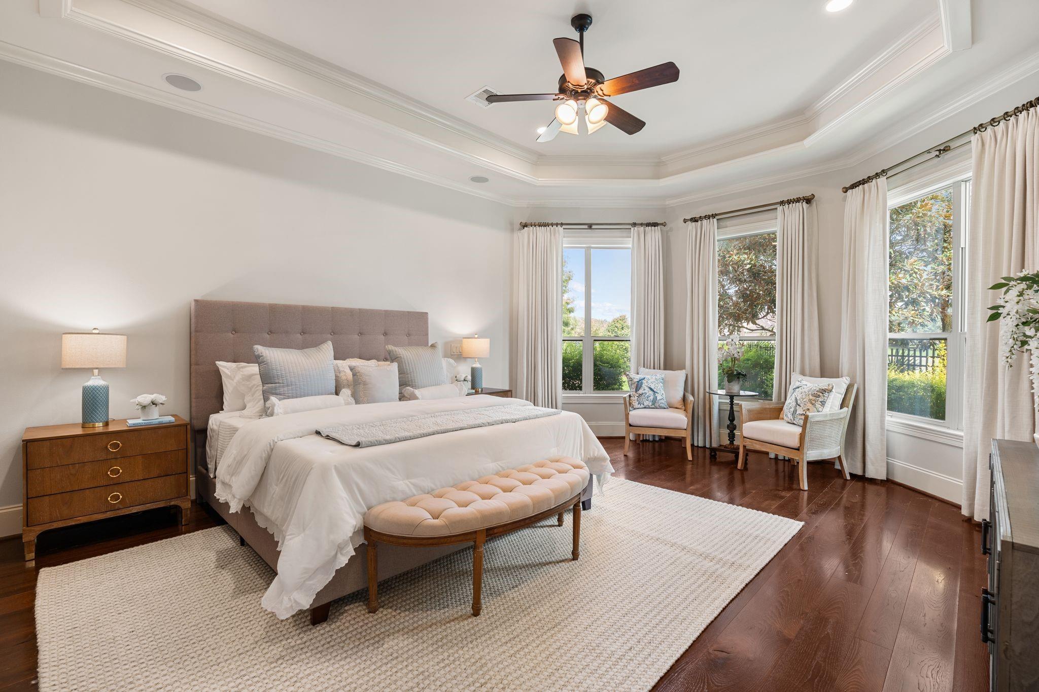 Staged primary bedroom with a king bed, tray ceiling, dark wood floors, and a bright window-side sitting area.