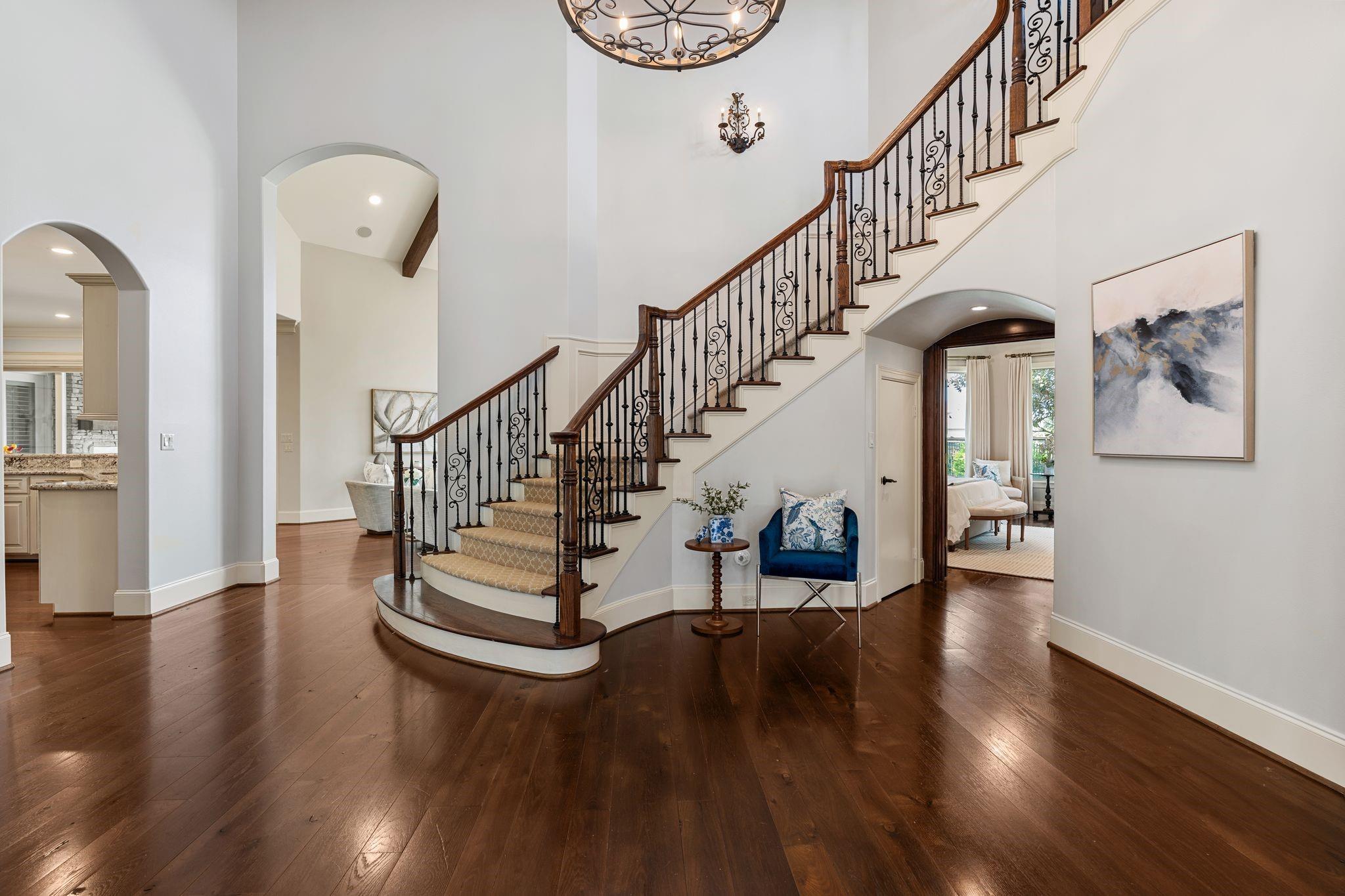 Foyer with a sweeping curved staircase, ornate iron balusters, hardwood floors, and a blue accent chair.