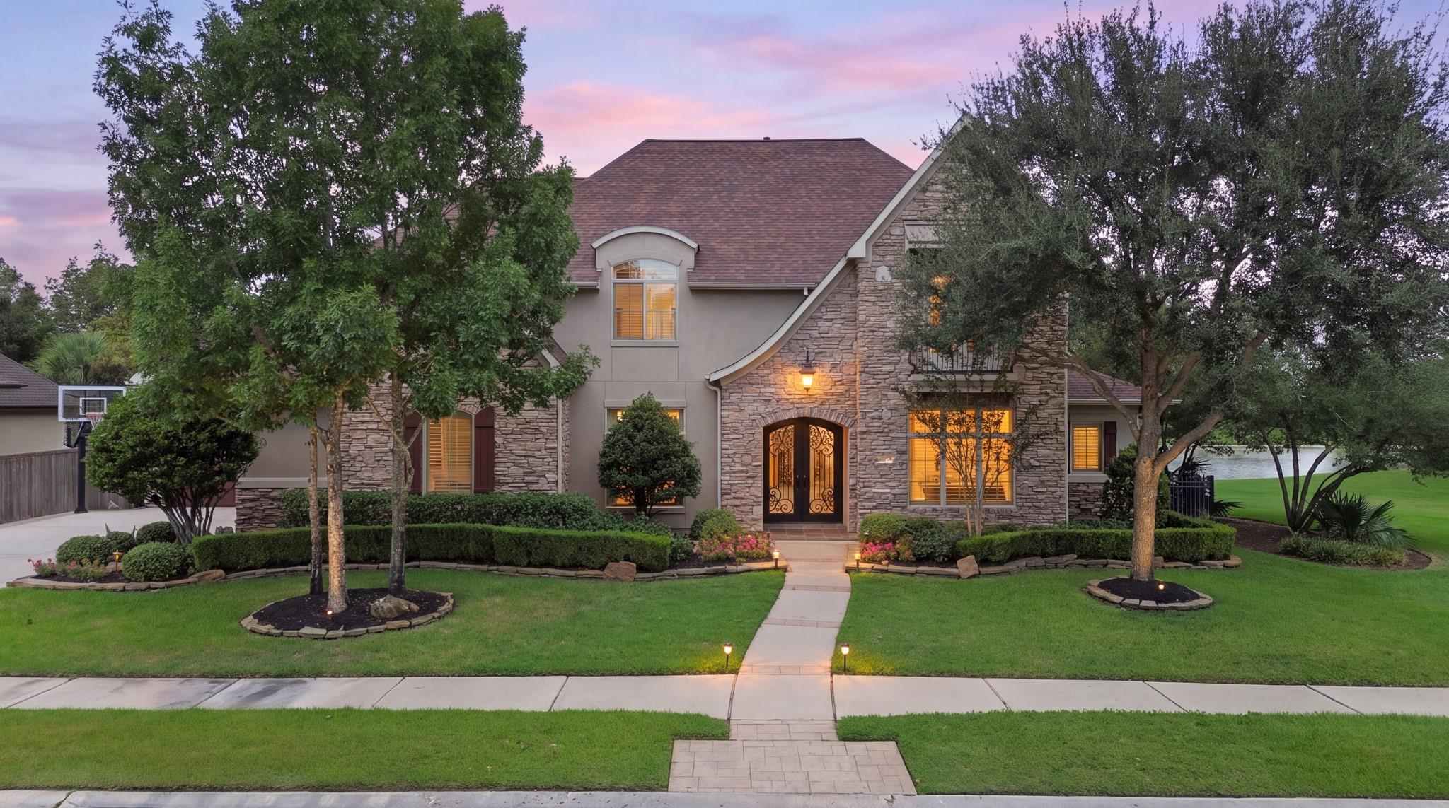 Two-story stone-and-stucco home at dusk, with glowing windows, mature trees, and a landscaped front lawn.