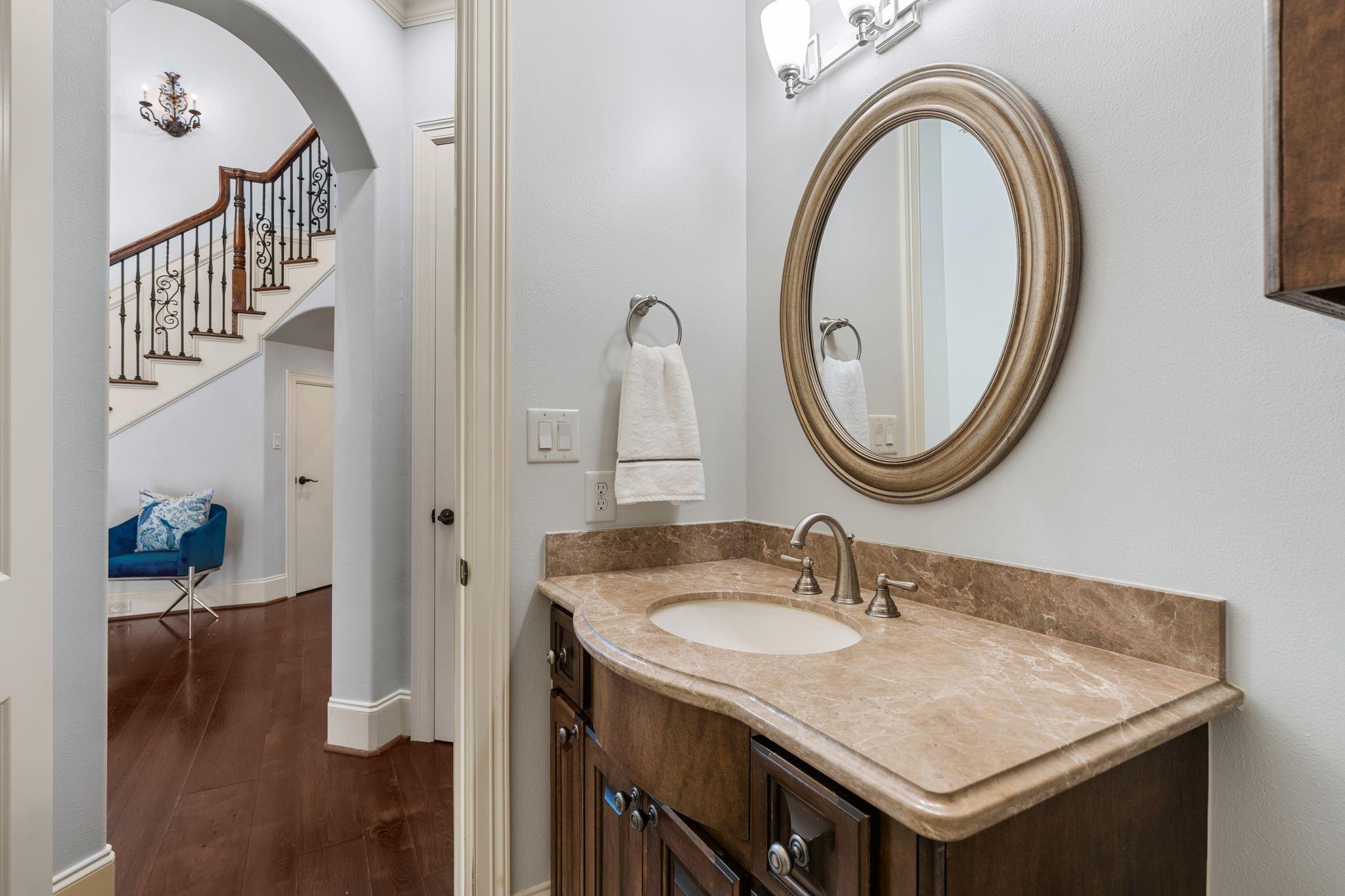 Bathroom vanity with a tan stone counter and oval mirror, overlooking an arched doorway and staircase.