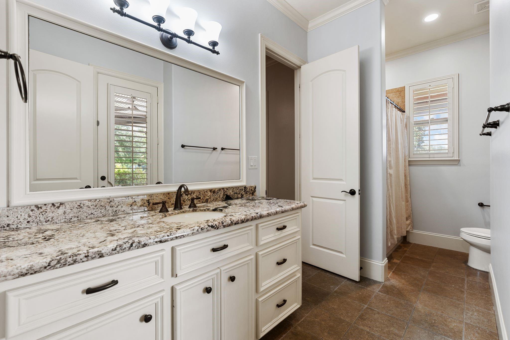 Bathroom with a granite-topped vanity, large mirror, shuttered window, and tiled floor.