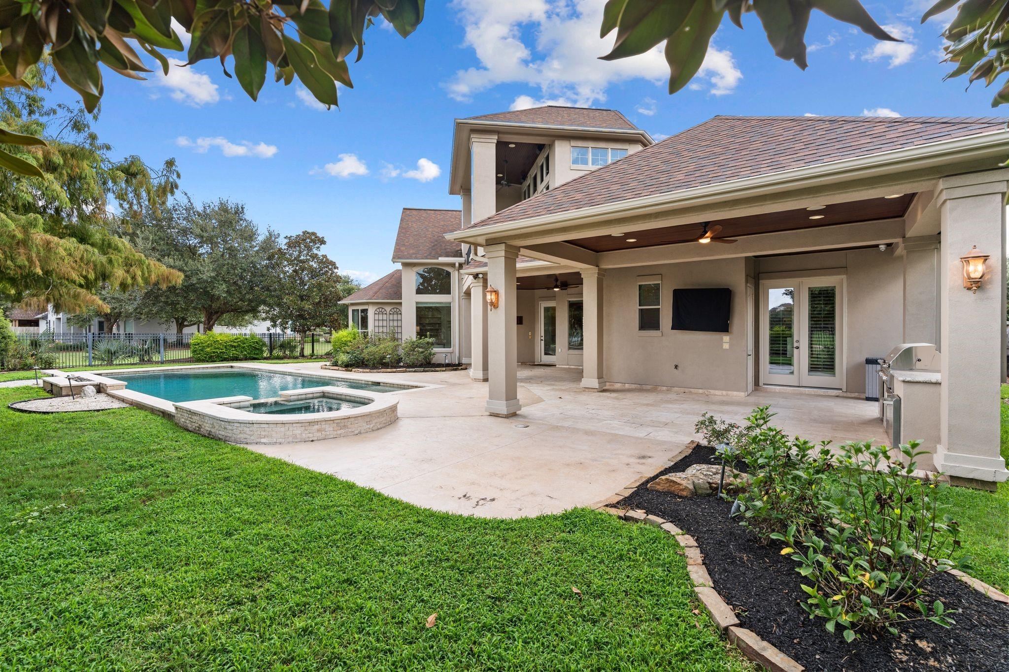 Home’s covered backyard patio beside a swimming pool and raised spa, with an outdoor grill and ceiling fans.