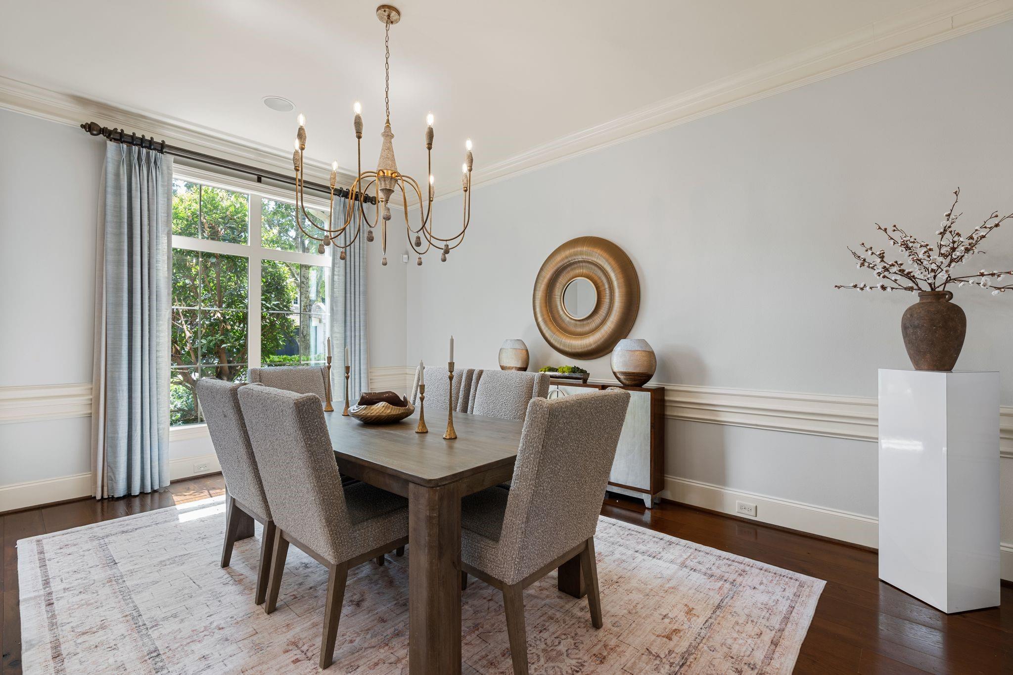 Dining room with a dark wood table, upholstered chairs, brass chandelier, and large curtained window.