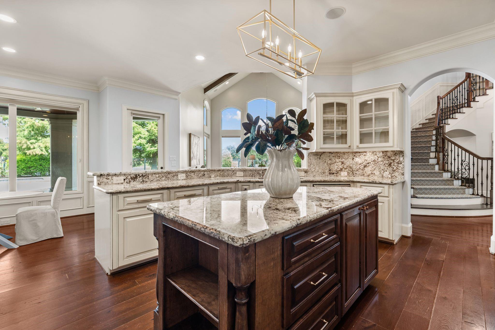 Kitchen with a large granite-topped island, white cabinetry, tall arched windows, and a carpeted staircase.