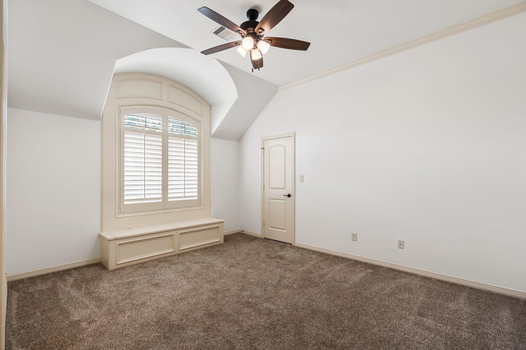 Empty bedroom with a built-in window seat, arched shuttered window, and ceiling fan.