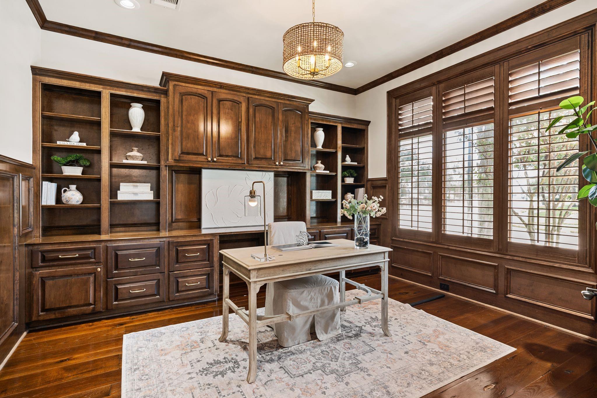 Home office with dark wood built-ins, plantation shutters, and a light desk on a patterned rug.
