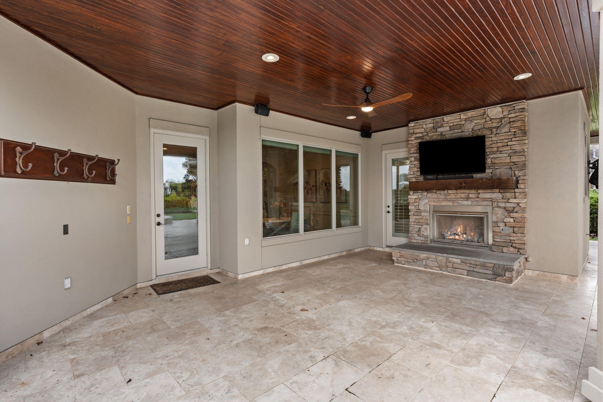 Covered patio with a stone fireplace, television, tiled floor and dark wood ceiling.