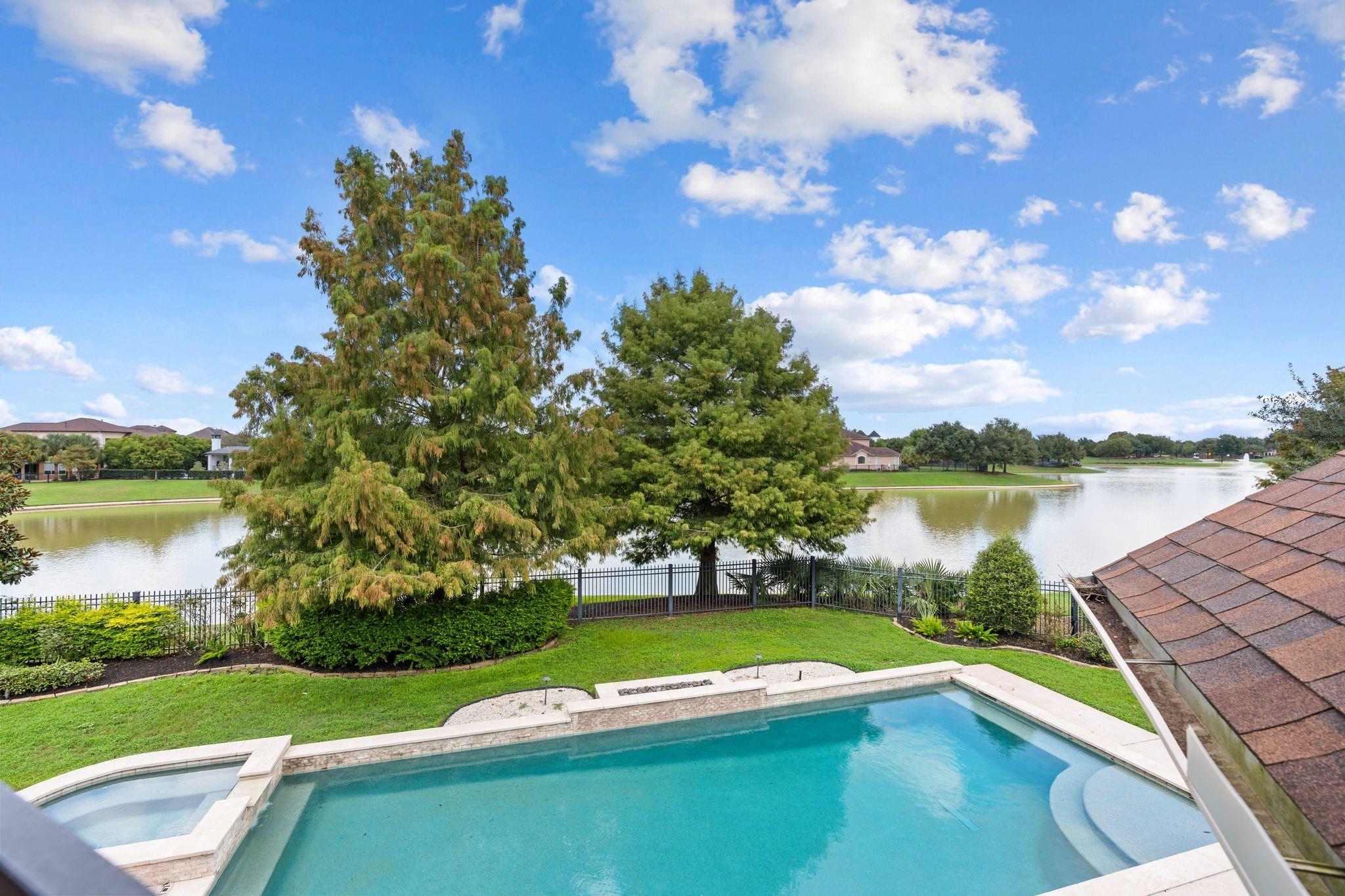 Backyard pool and spa overlooking a lake, with mature trees along the fenced lawn.