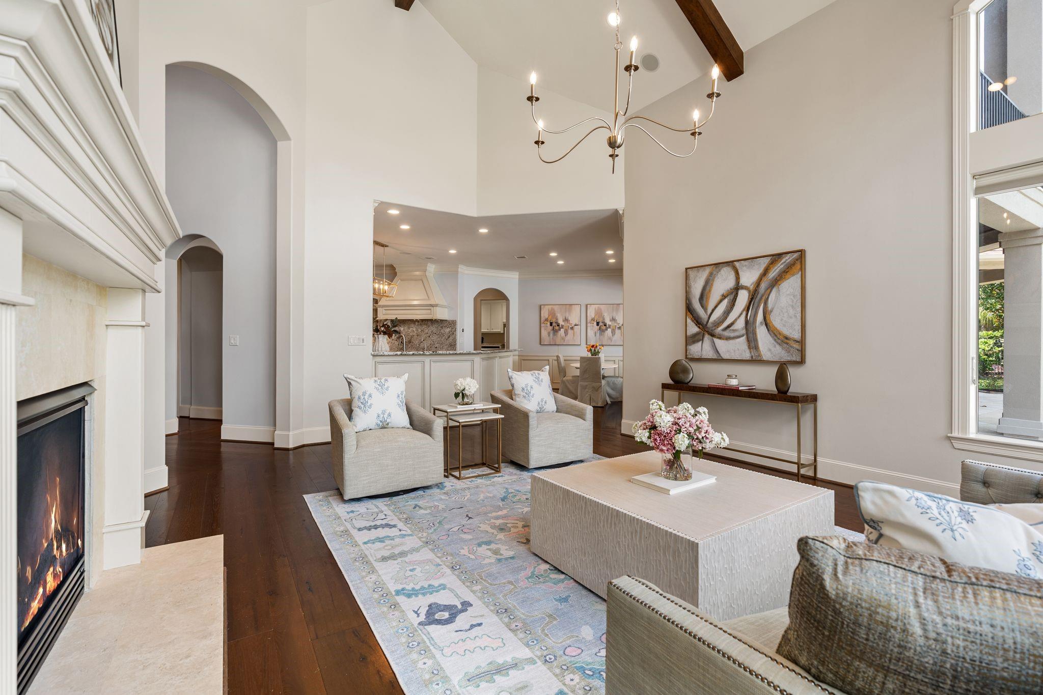 Living room with a lit fireplace, vaulted ceiling, exposed beam, and open connection to the kitchen and dining area.