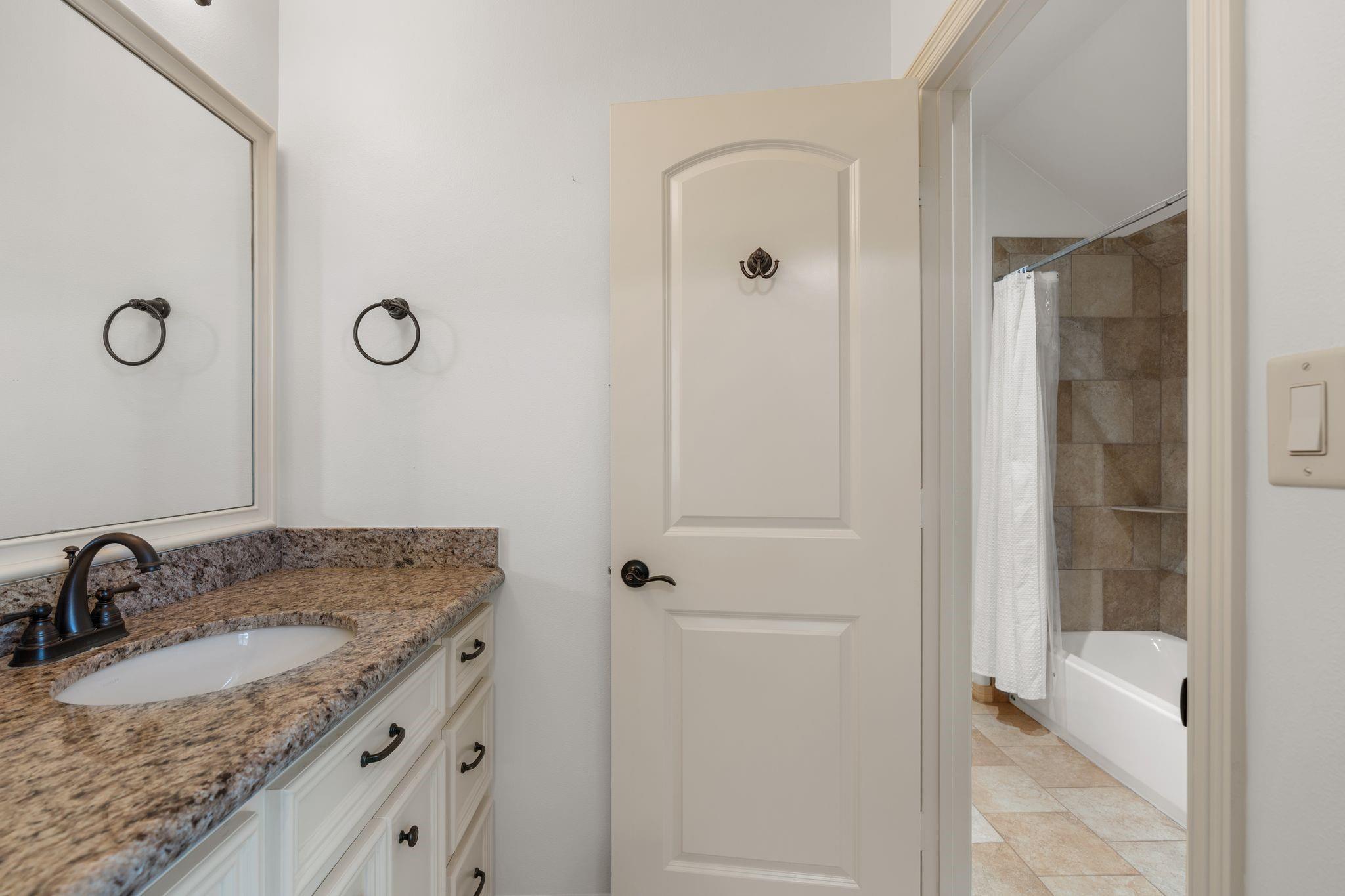 Bathroom vanity with granite countertop and mirror beside a tan-tiled tub and shower.