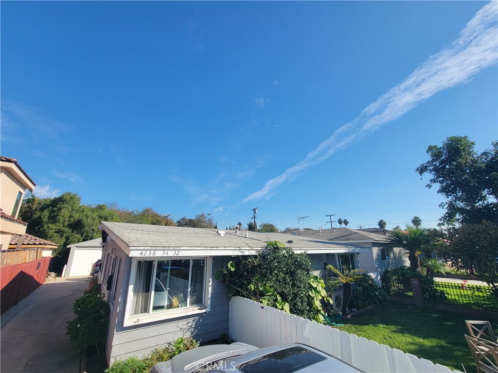 Single-story gray home with a fenced garden and parked car beneath a broad blue sky.