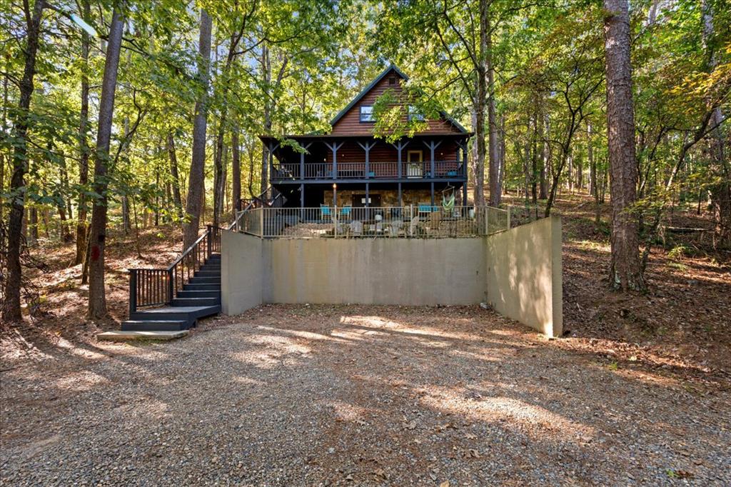 Two-story wooded cabin with a covered balcony, raised stone base, and broad gravel driveway.