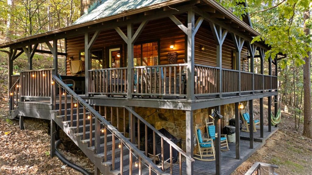 Raised log cabin with a covered wraparound porch and staircase in a wooded setting.