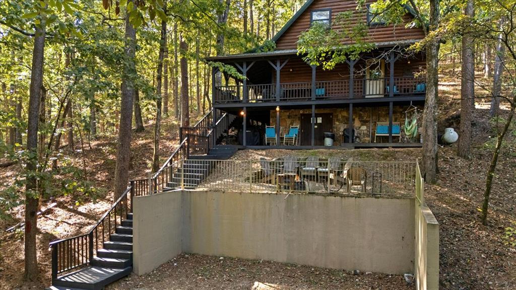 Log cabin among trees with covered decks, outdoor seating, and a staircase to a fenced patio.