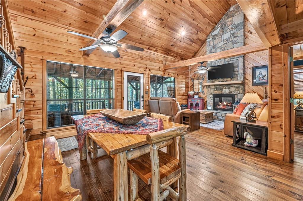 Log-cabin living and dining room with vaulted wood ceiling, stone fireplace, and windows overlooking trees.