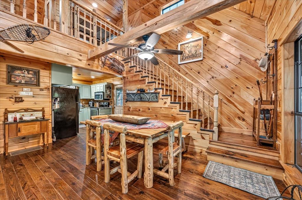 Rustic cabin dining area with a log table, open kitchen, and staircase to a loft.