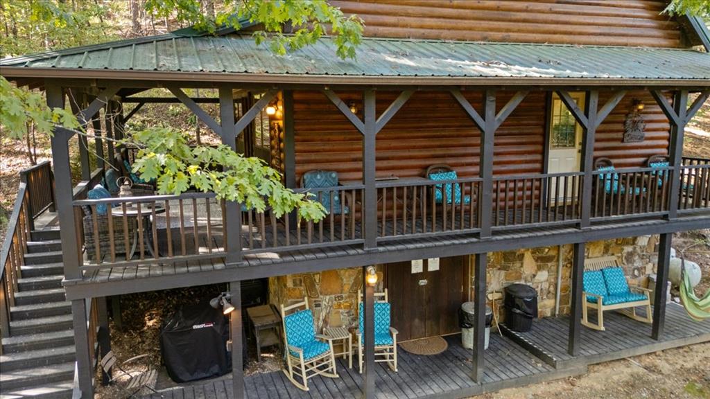 Log cabin with two levels of covered decks, outdoor seating, and stairs among leafy trees.