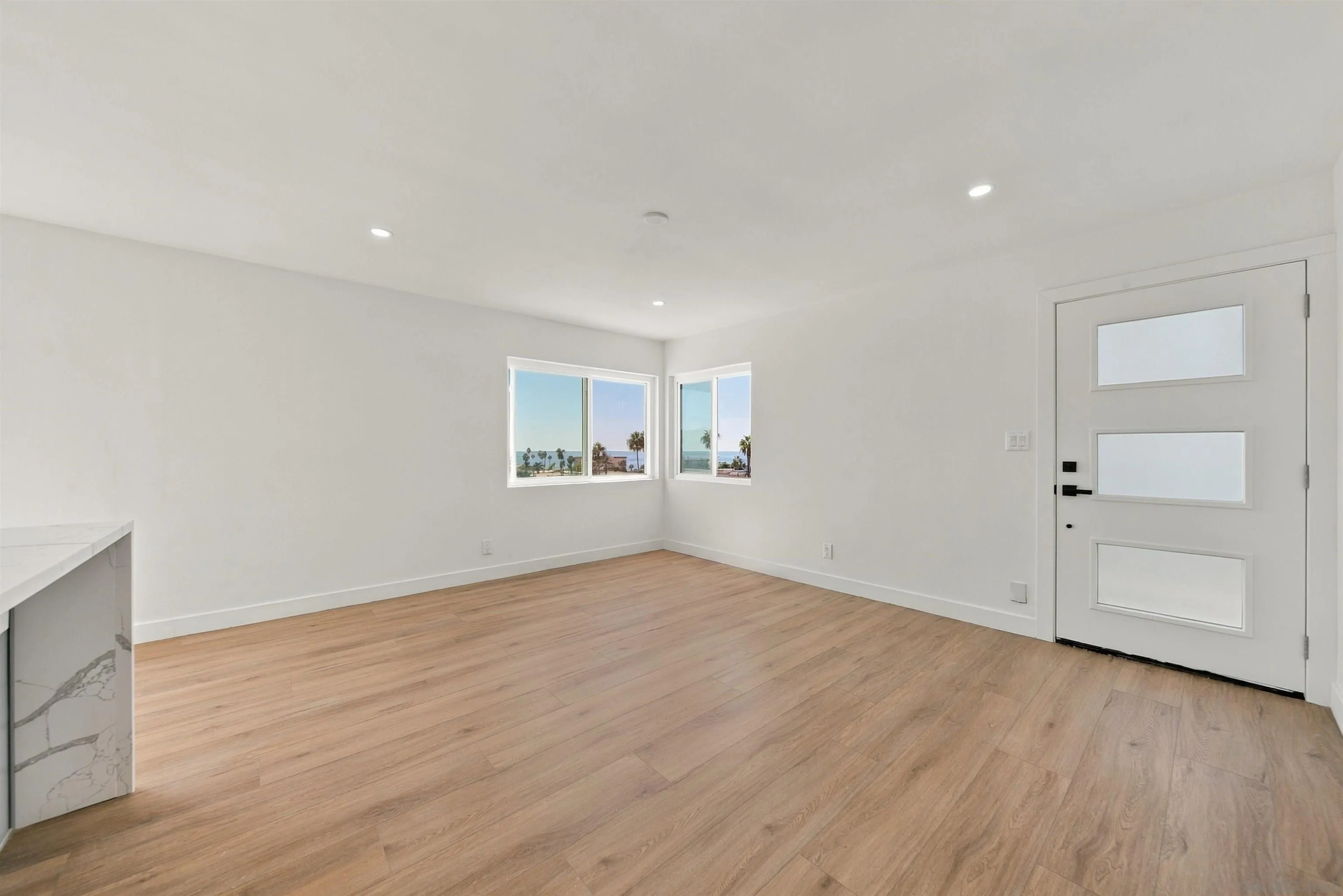 Vacant room with light wood floors, modern white entry door, and ocean views through corner windows.