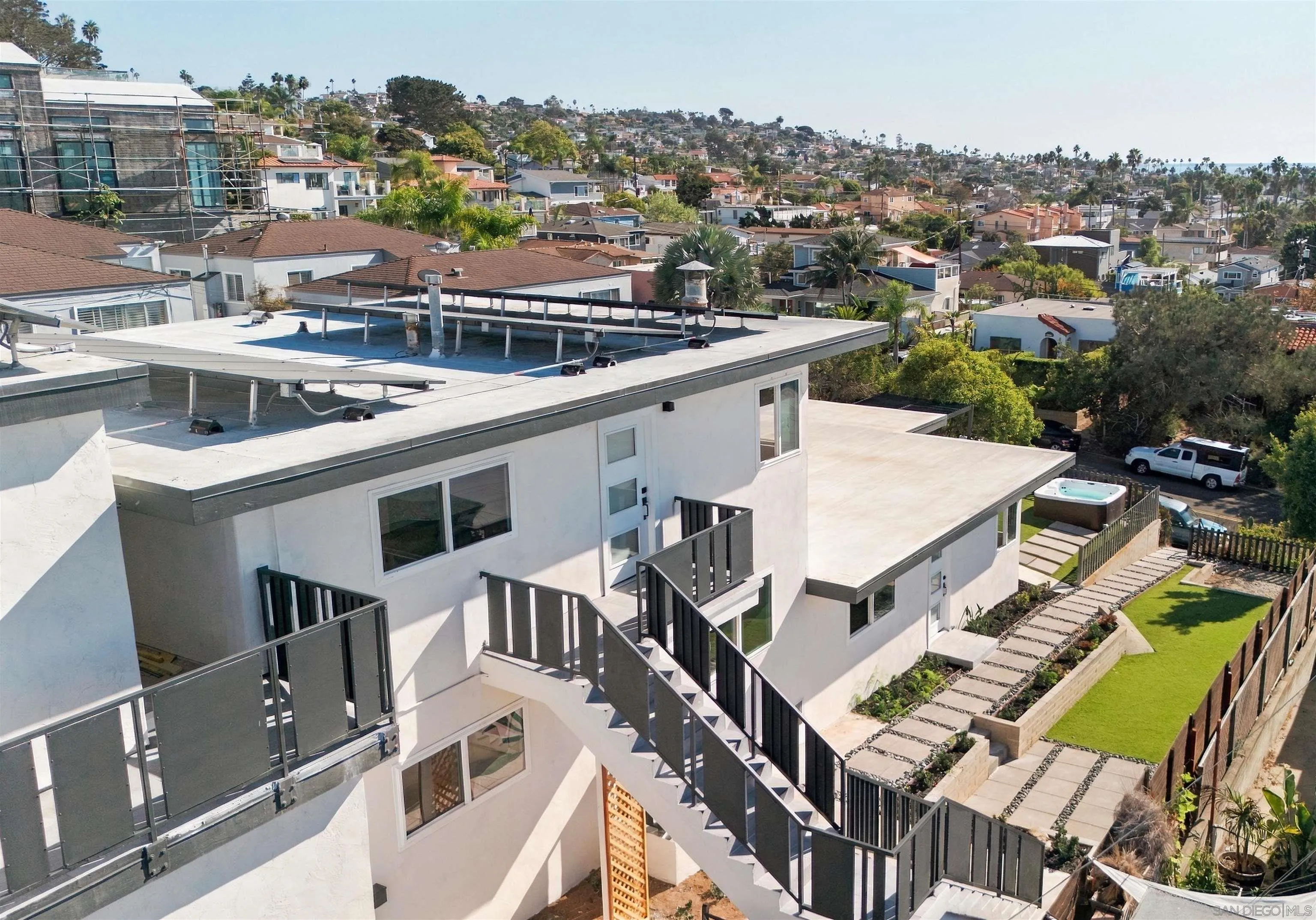 Modern white multi-level property with rooftop solar panels, exterior stairs, landscaped lawn, and hot tub.