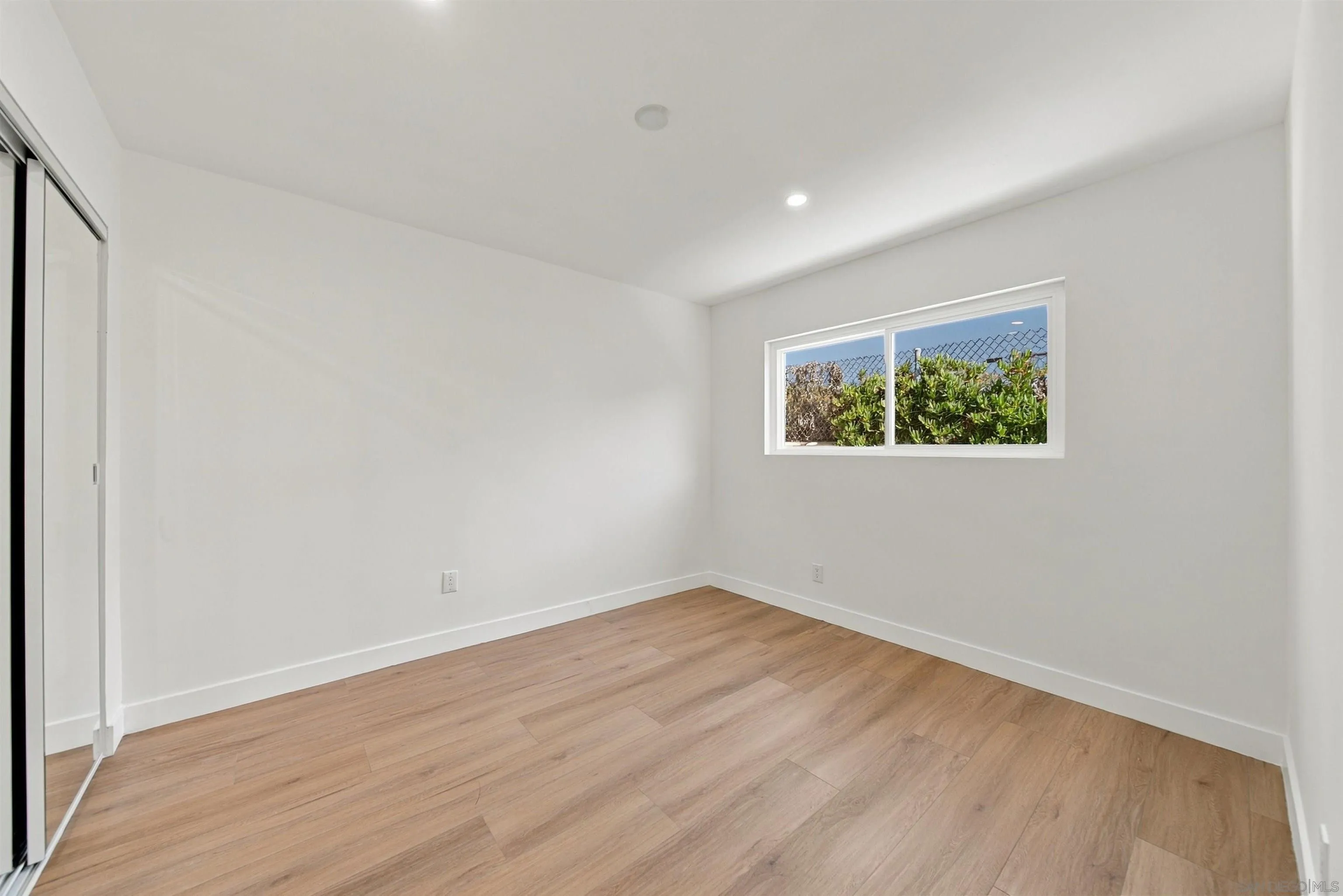 Empty bedroom with wood flooring, mirrored closet doors, and a window overlooking greenery.
