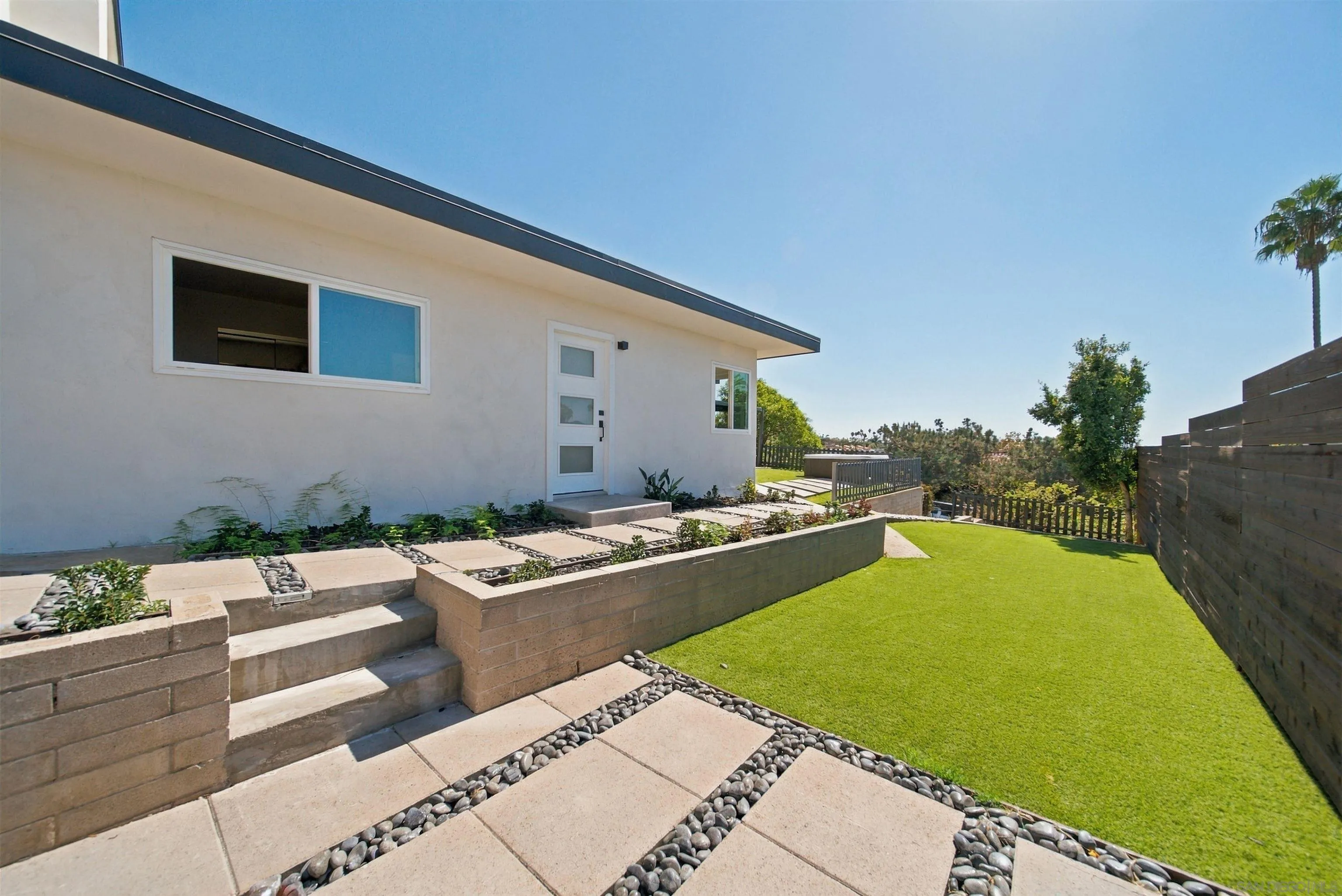 Modern white house beside a landscaped backyard with a stone path, raised beds, and bright green lawn.