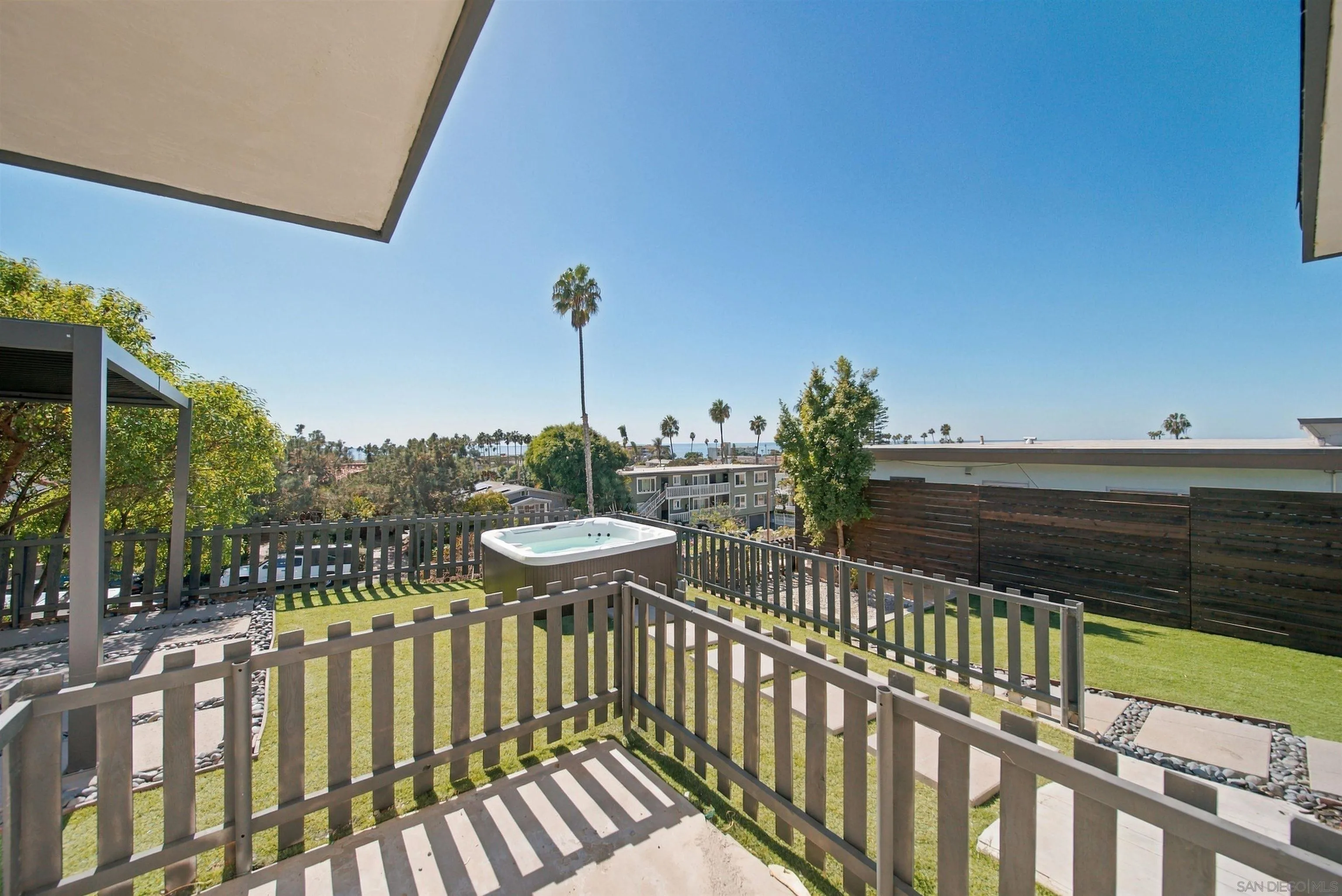 Outdoor patio with a hot tub and fenced lawn, with palm trees and a glimpse of ocean beyond.