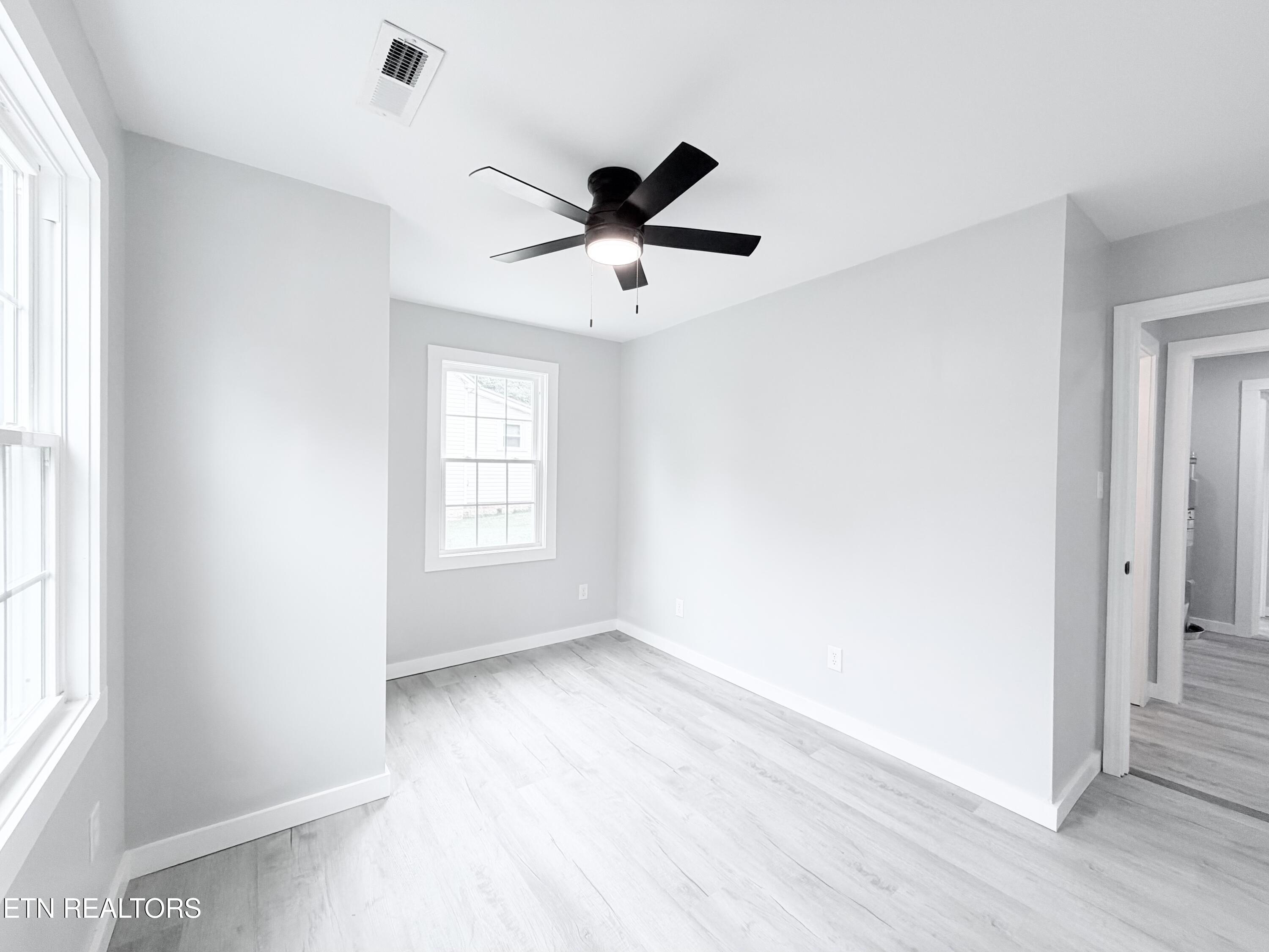 Vacant room with pale walls, light flooring, two windows, and a black ceiling fan.