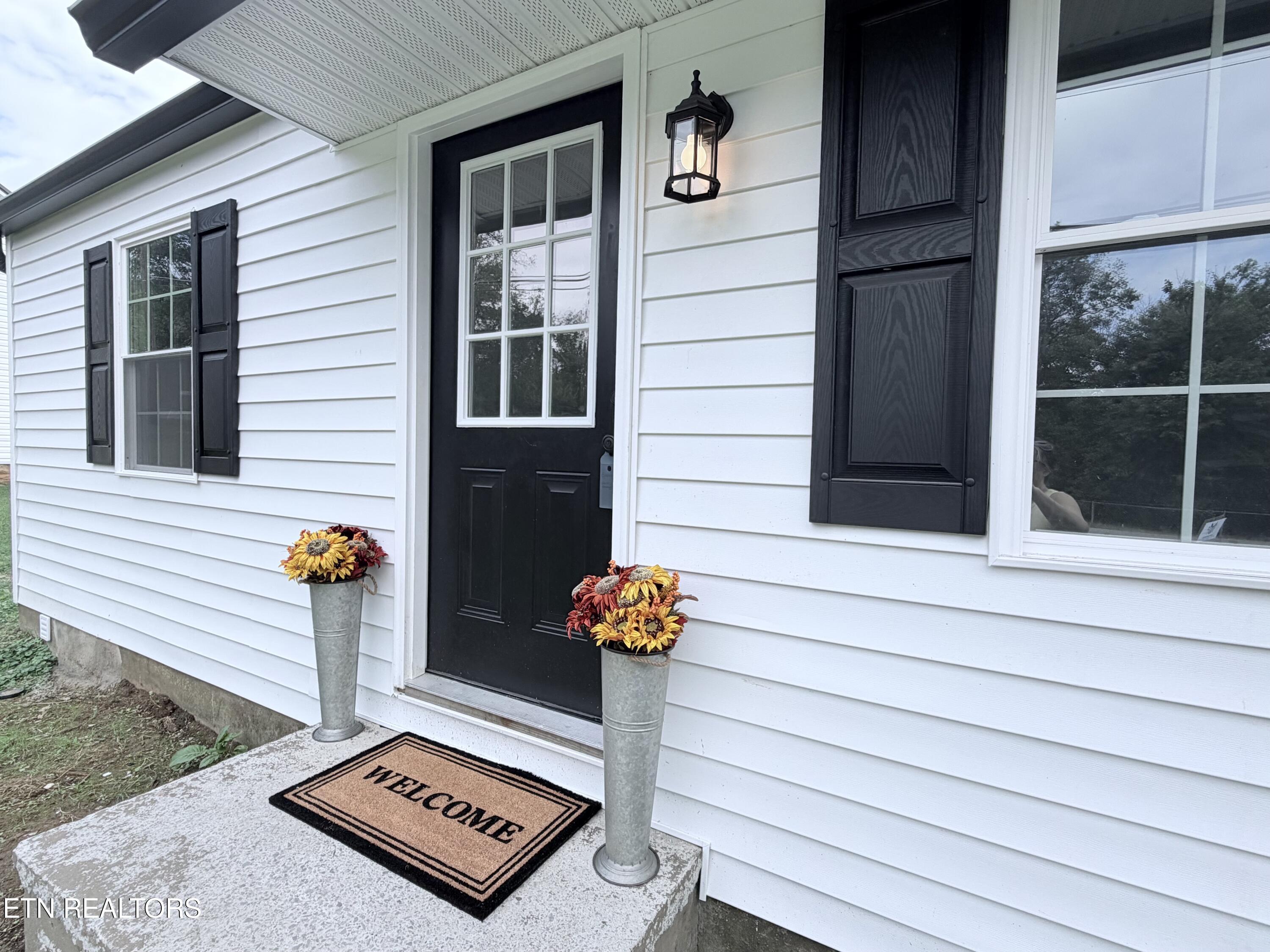 Home’s black front door framed by white siding, flower planters, and a welcome mat.