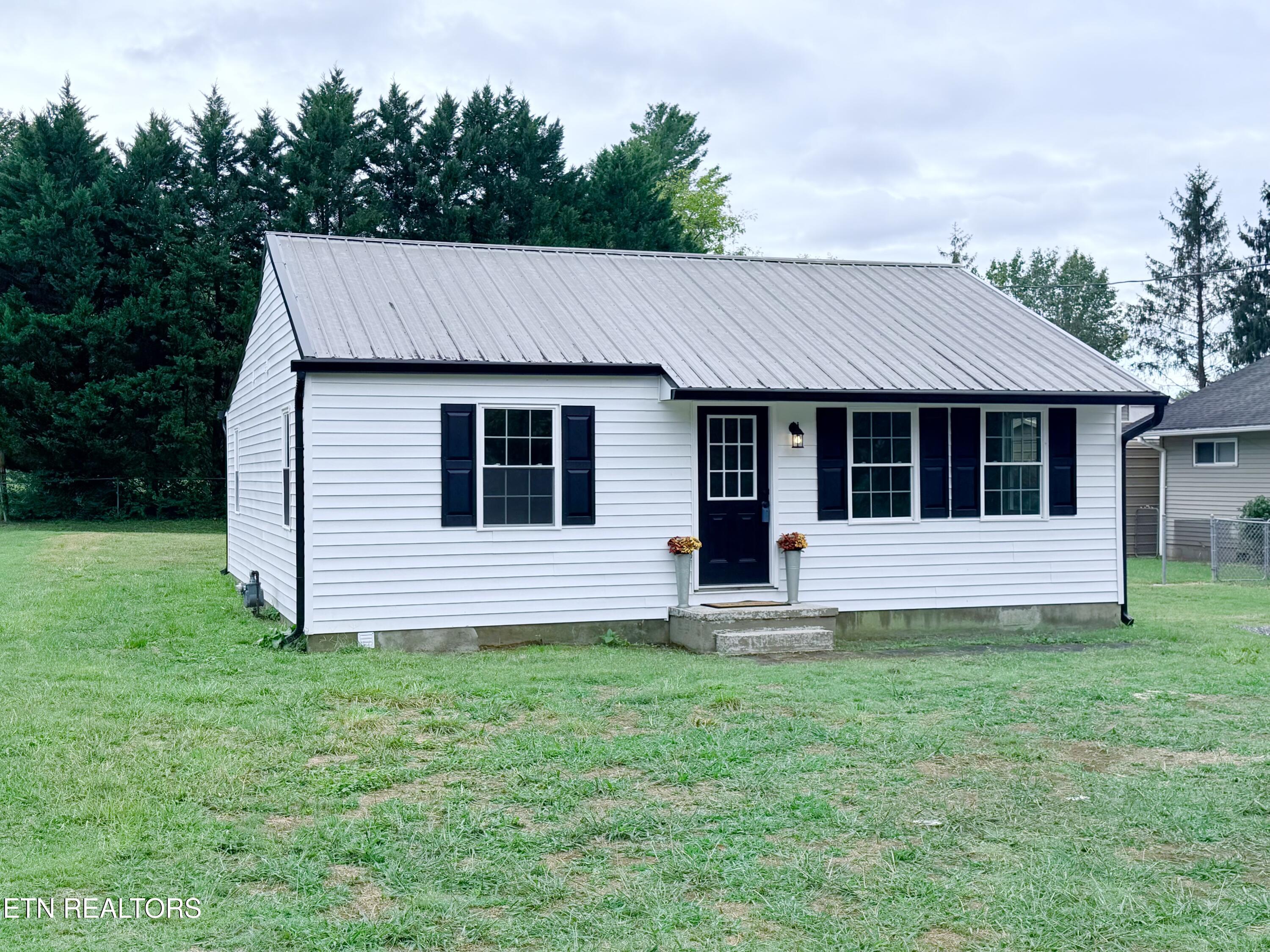 White house with a light metal roof, navy shutters, and a grassy front lawn.