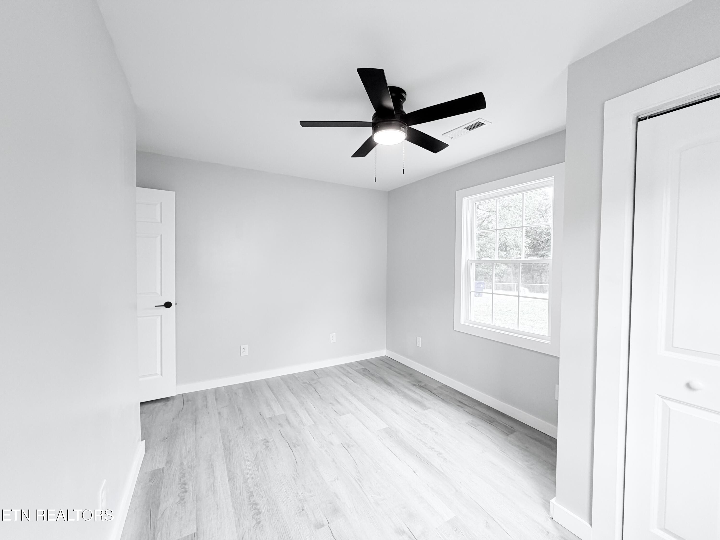 Empty bedroom with pale plank flooring, a black ceiling fan, and a window overlooking the yard.