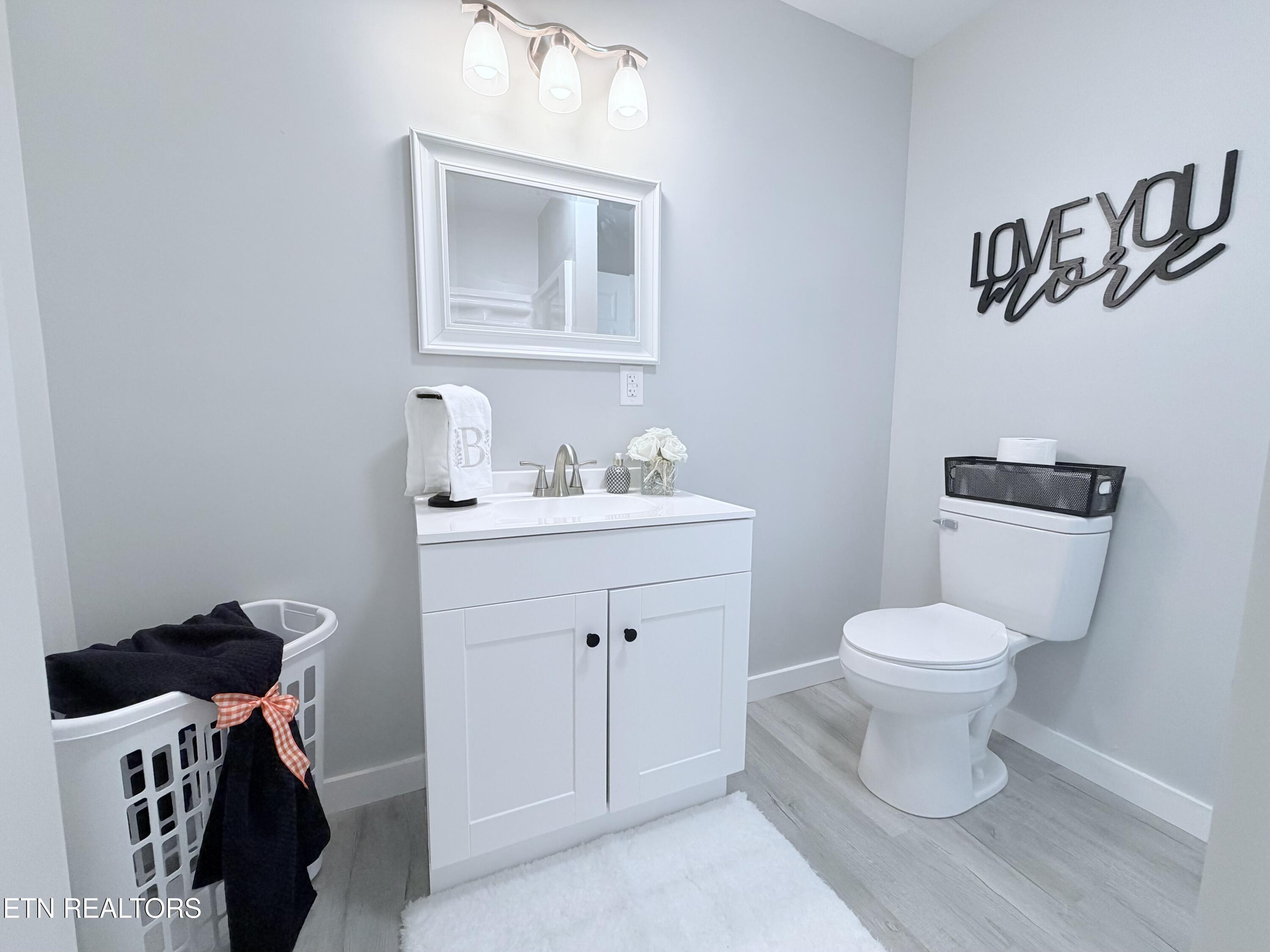 Staged bathroom with a white vanity, framed mirror, toilet, and laundry basket with an orange gingham bow.