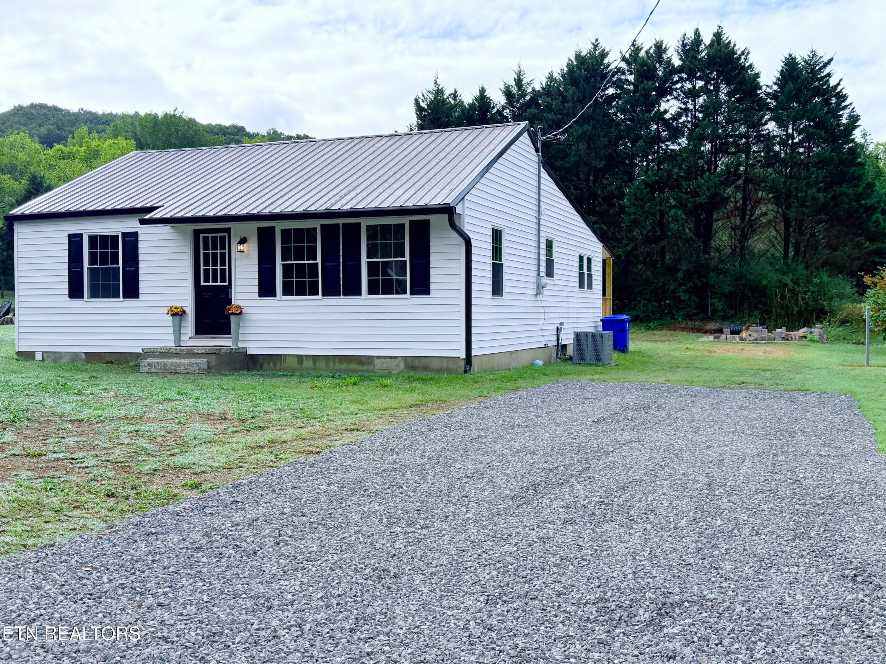 White single-story house with dark shutters, metal roof, and broad gravel driveway beside woods.