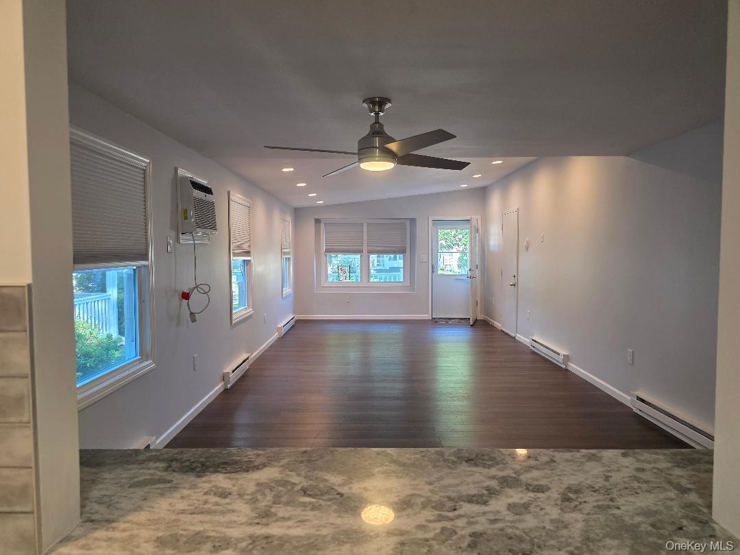 Vacant living room with dark wood flooring, ceiling fan, and windows with blinds.