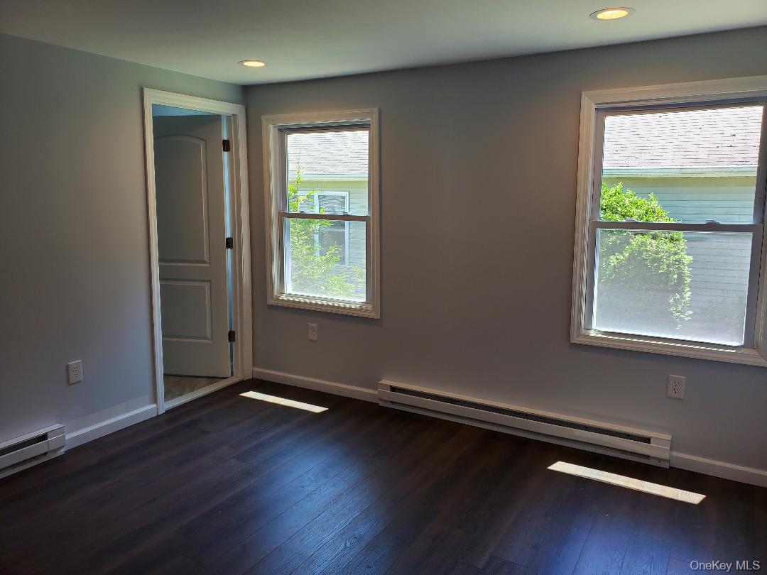Empty room with gray walls, dark wood flooring, two windows, and baseboard heaters.