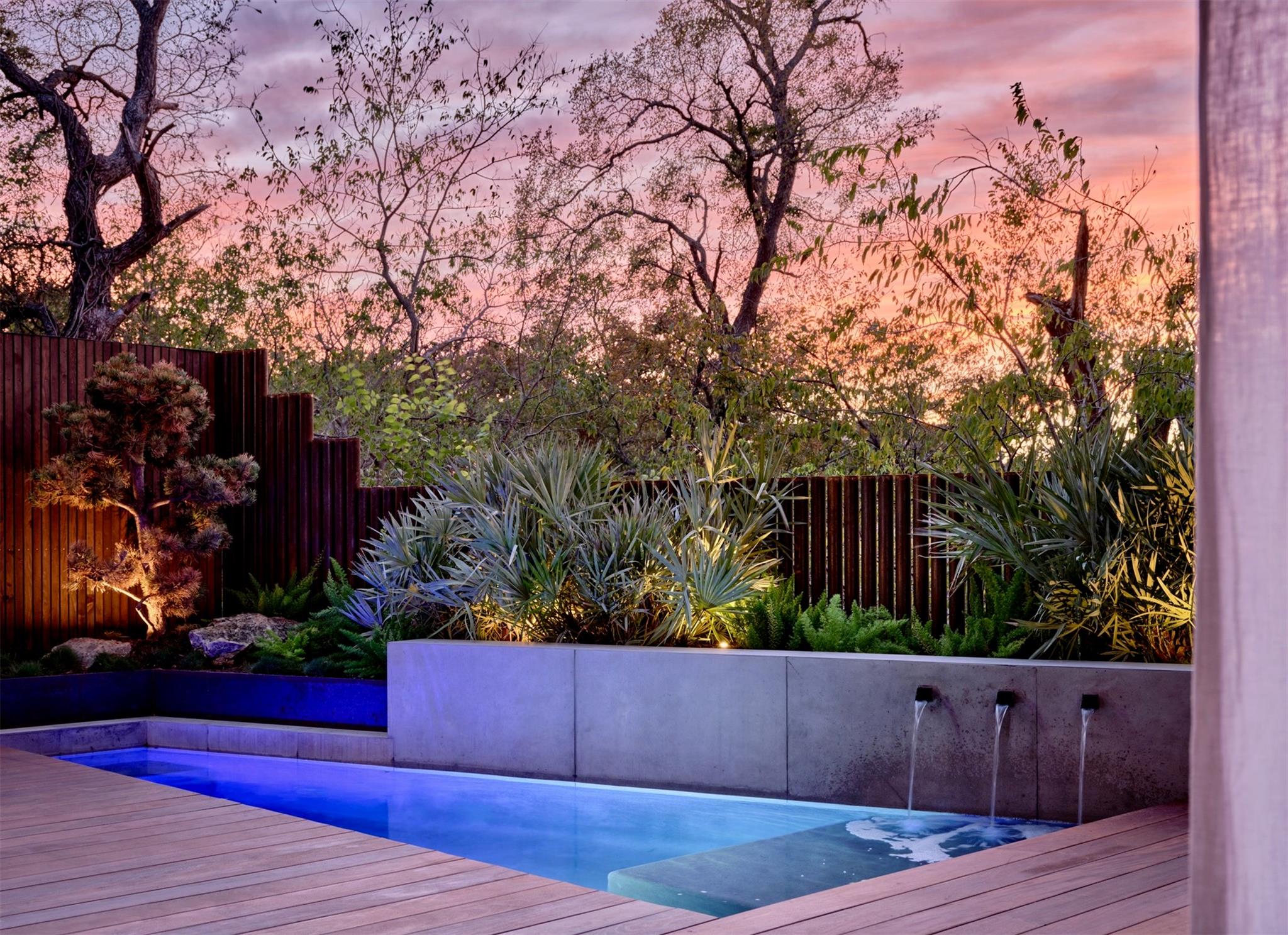 Backyard pool with three wall fountains, tropical landscaping, and a wood deck at dusk.