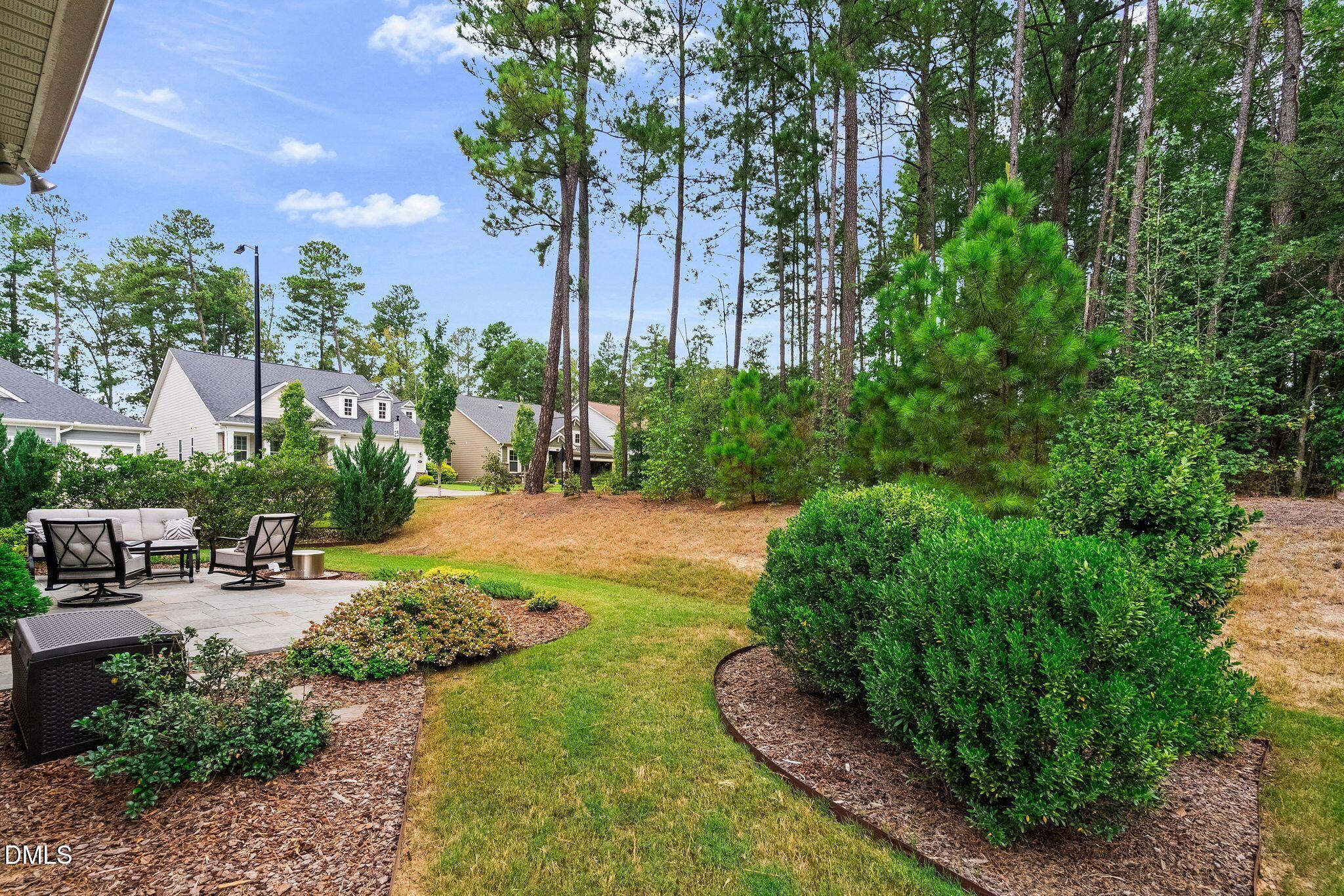 Backyard patio with outdoor seating, landscaped lawn, shrubs, and tall pine trees.
