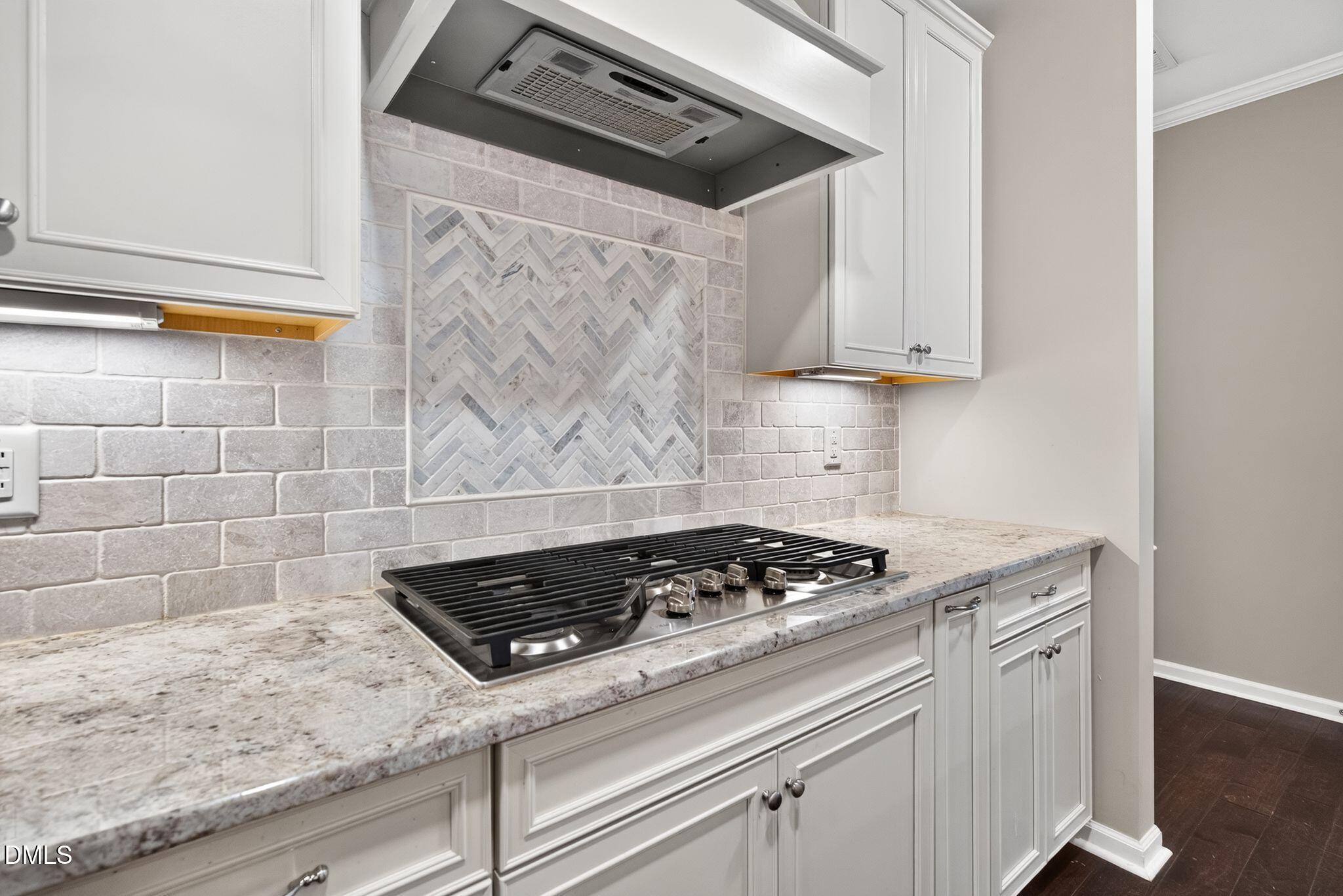 Kitchen gas cooktop beneath a gray-and-white herringbone backsplash, with white cabinets and stone counters.