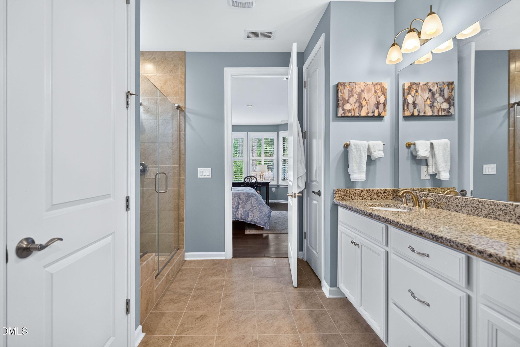 Bathroom with a glass-enclosed tile shower, granite double vanity, and doorway to a bedroom.
