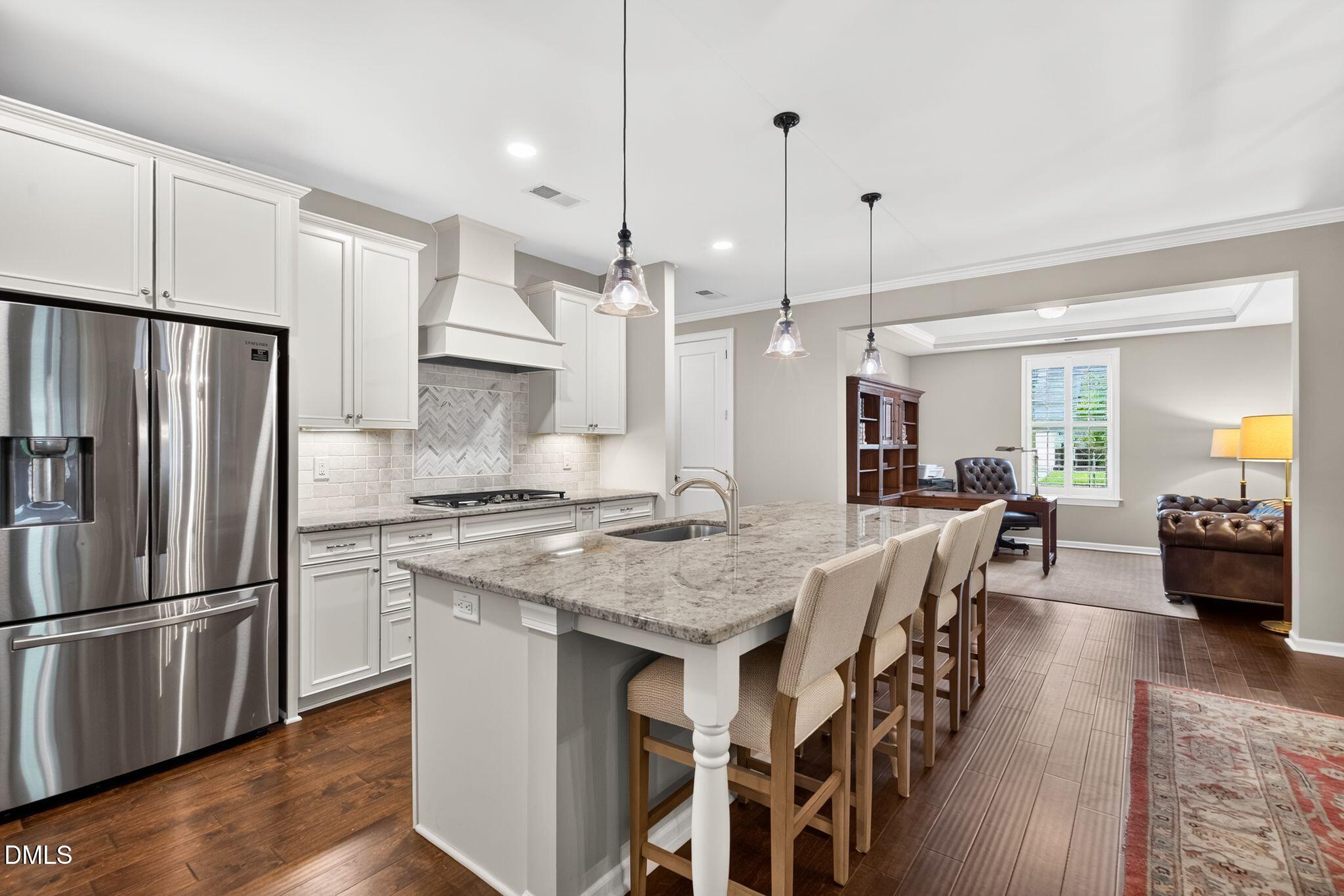 Bright white kitchen with a granite island and bar seating, opening to a furnished home office and living area.