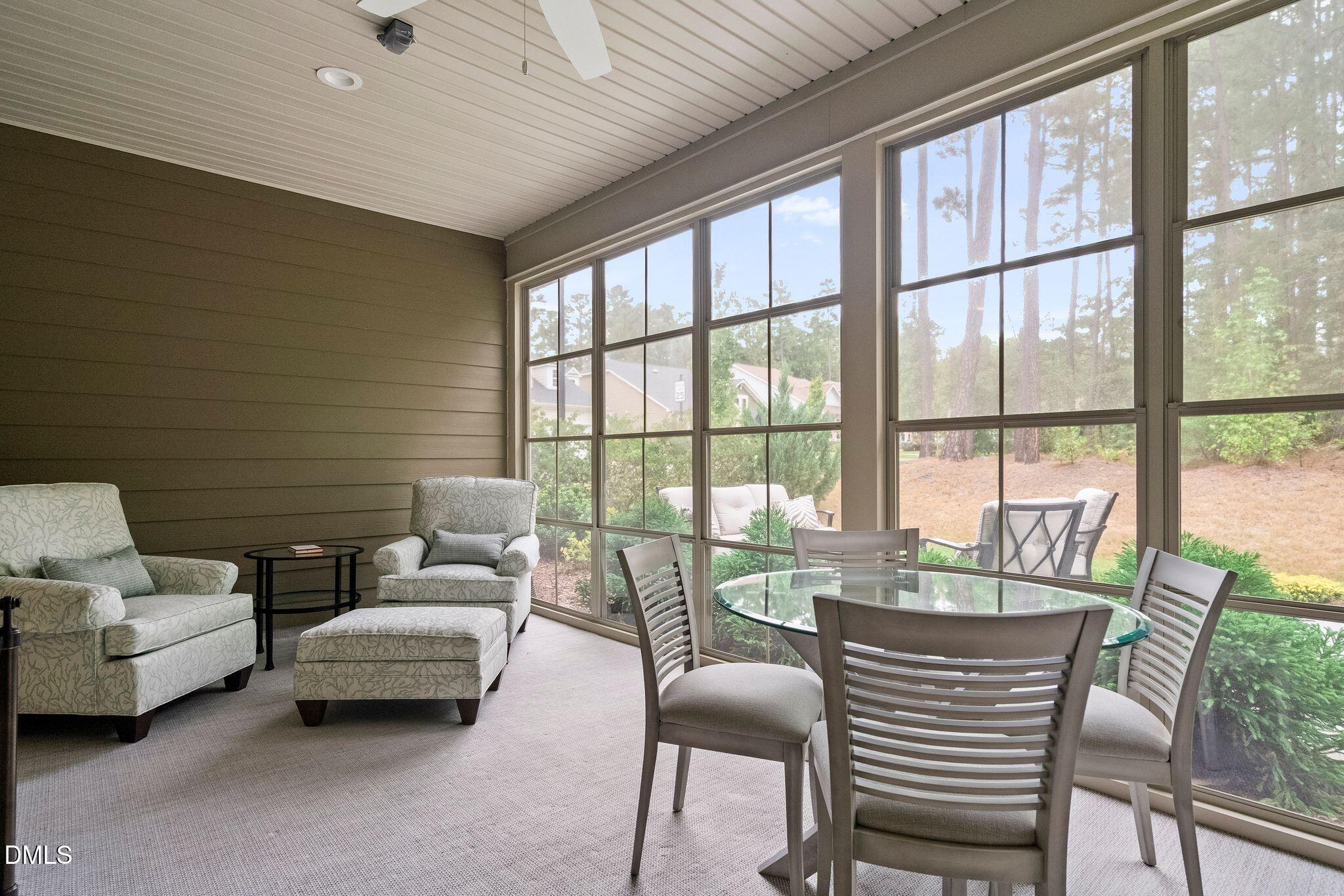 Furnished sunroom with a glass dining table, armchairs, and large windows overlooking trees.