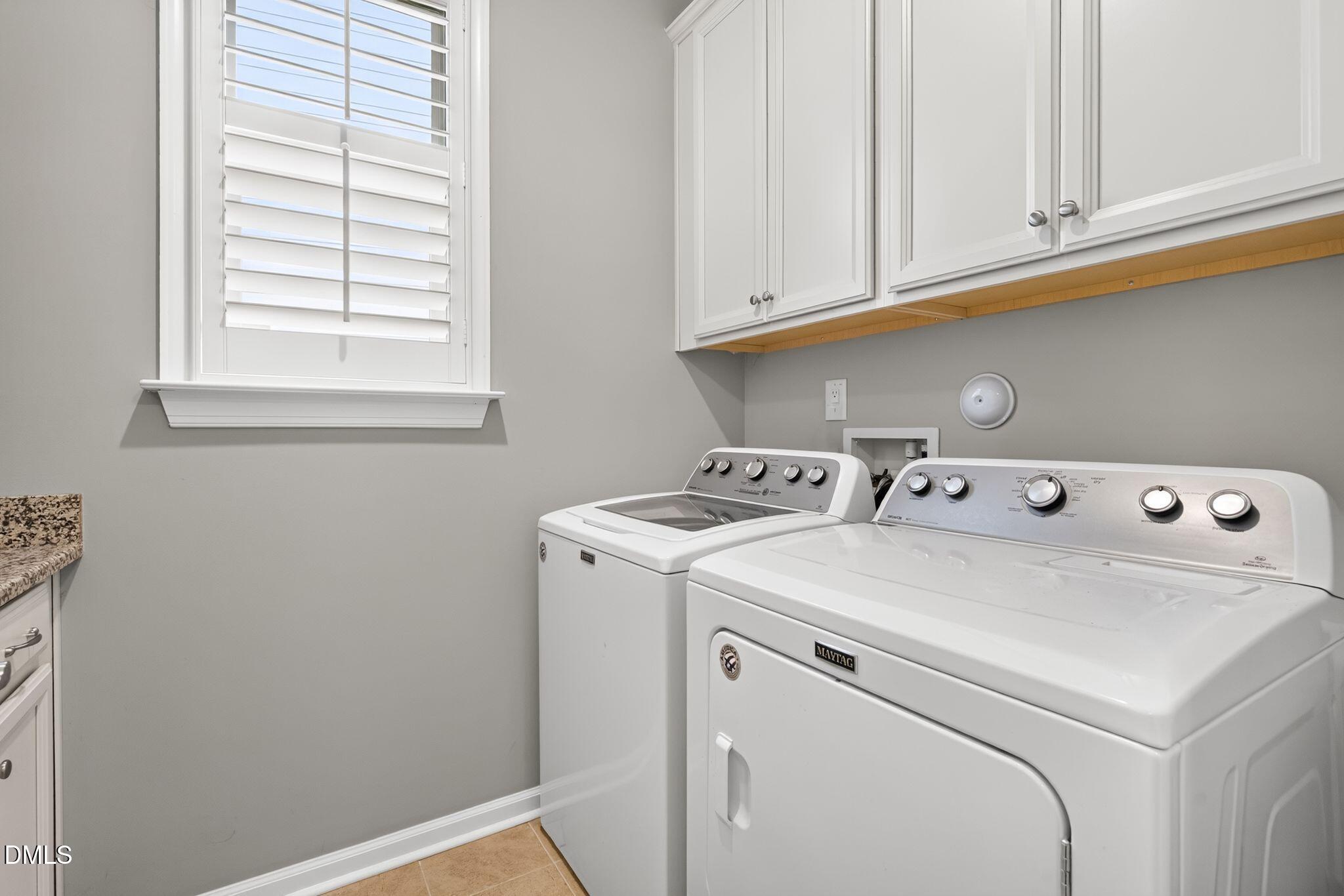 Laundry room with a white washer and dryer beneath cabinets beside a shuttered window.